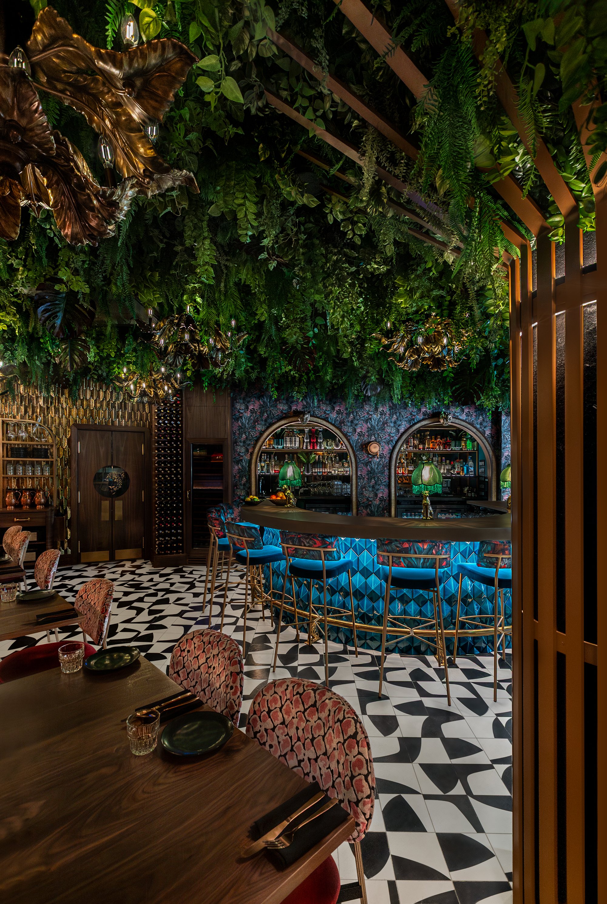 An upscale bar interior with a lush green leafy ceiling and decorative wall coverings, a curved bar with blue geometric pattern, patterned upholstered chairs, and a checkered black and white floor.