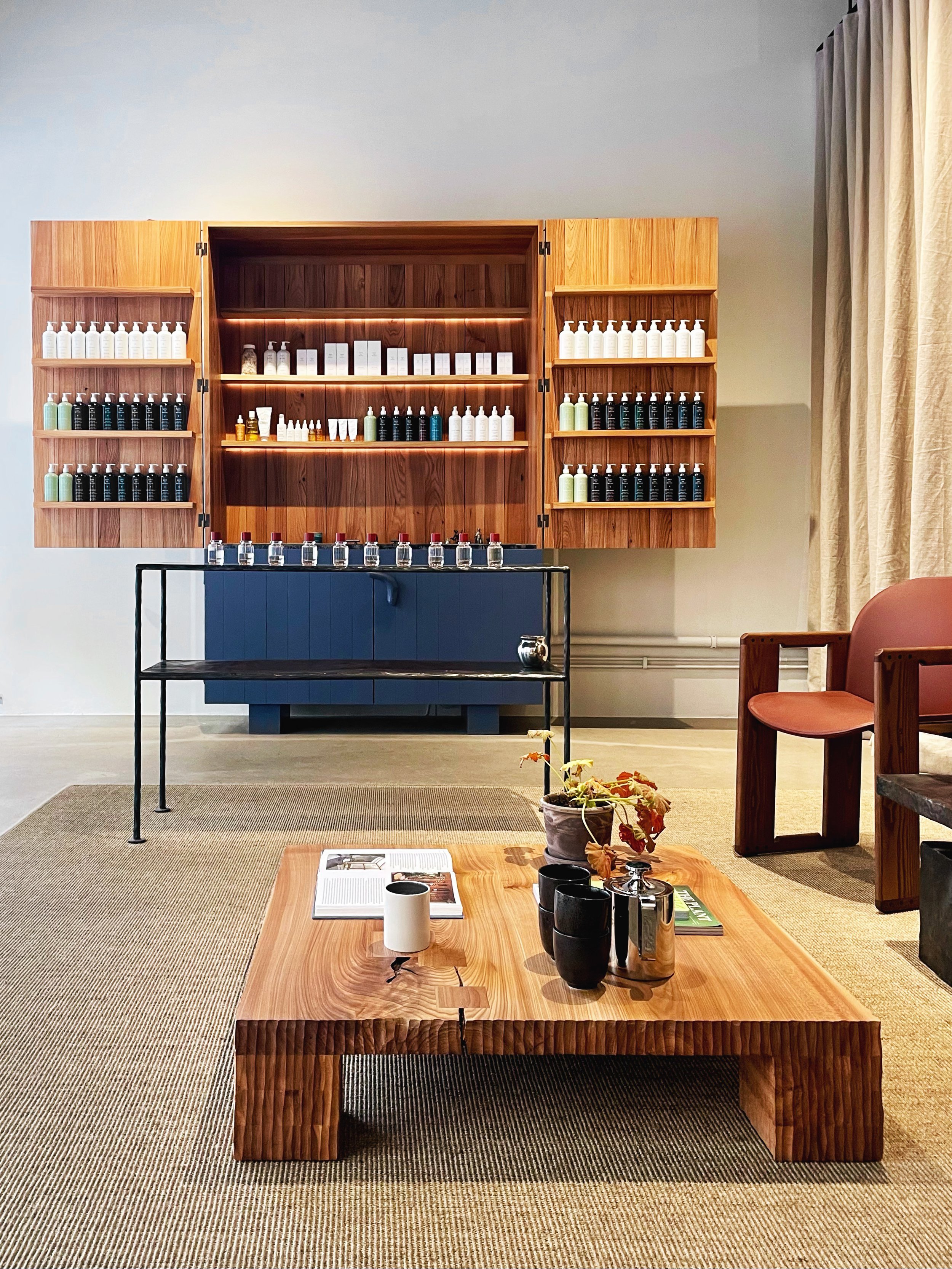 A display of aesthetic bottles and skincare products on wooden shelves in a minimalist room with a wooden table, a small potted plant, black cups, a magazine, and a cream mug on the table. The background features beige curtains and a neutral wall.