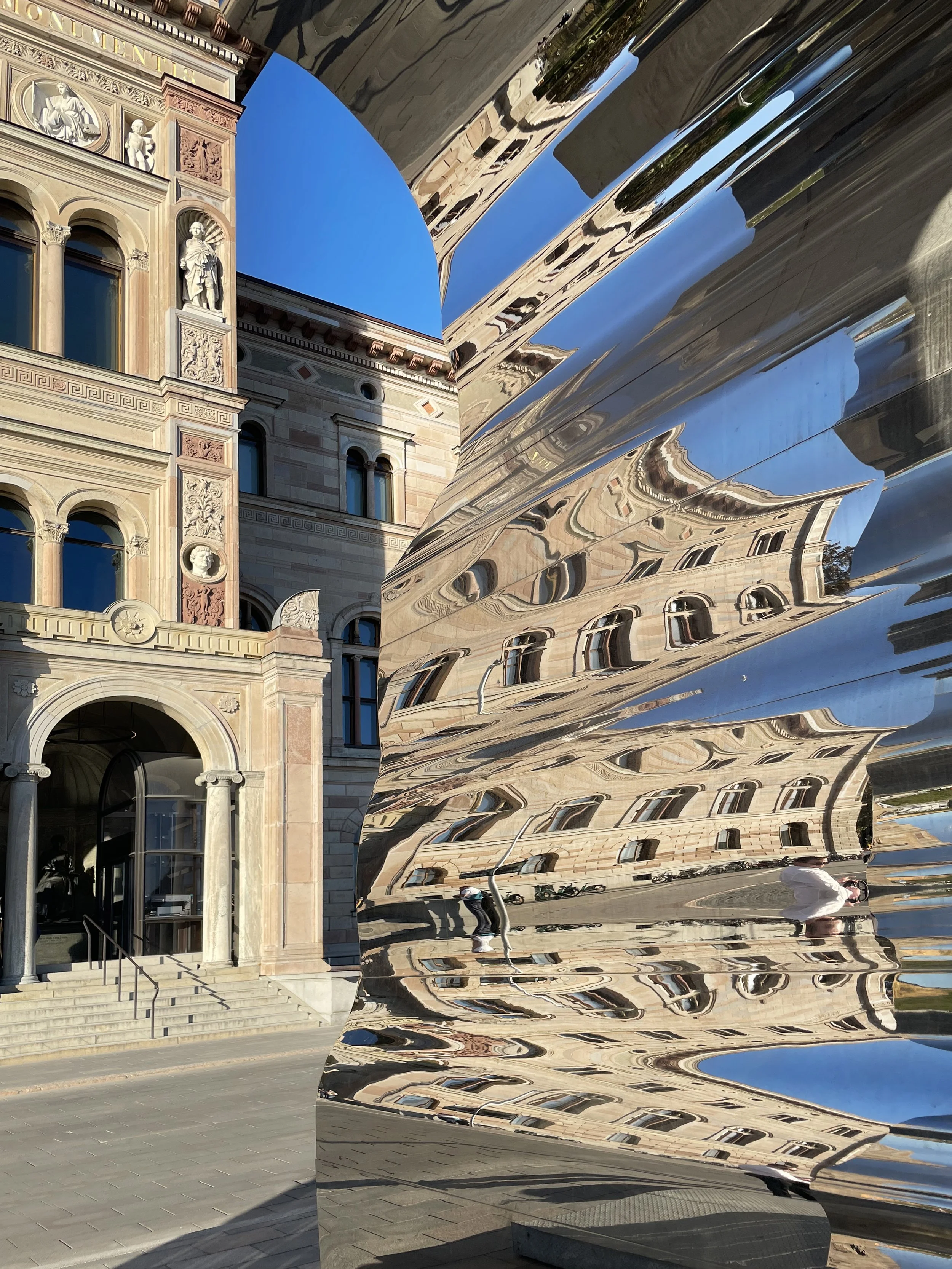 Reflection of a historic building in a large, twisted metallic sculpture, with people walking nearby under a clear blue sky.