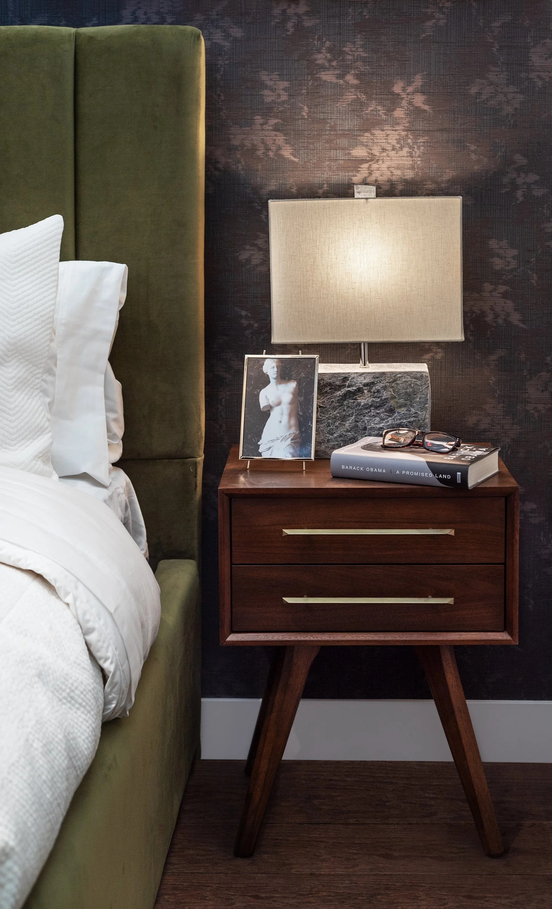 A bedside table next to a bed with white sheets and pillows. The table has two drawers with gold handles, a framed black-and-white art photo of a woman, a thick book titled 'Barack Obama | A Promised Land,' a pair of glasses, and a lamp with a stone