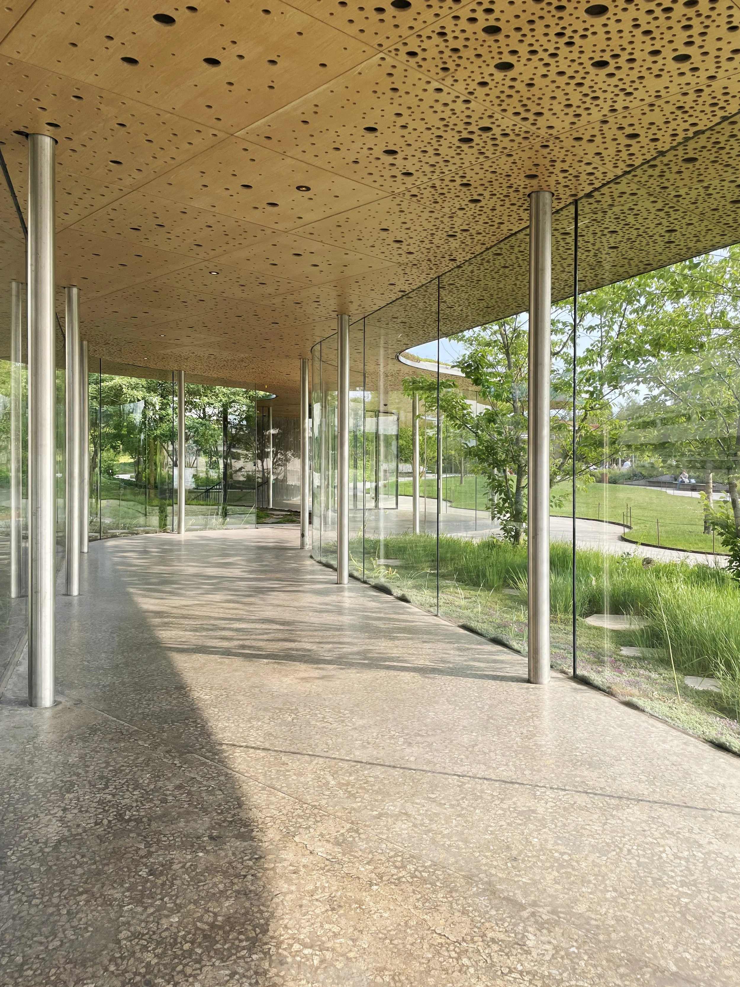 Modern building with a curved roof, glass walls, and metal supports, overlooking a landscaped green outdoor area with trees and grass.
