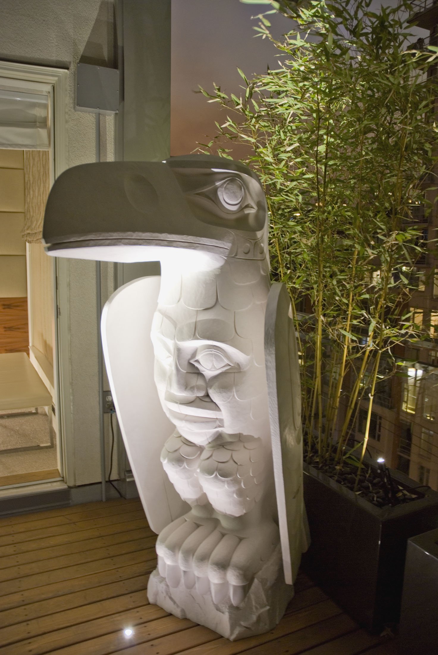 A large sculptural art piece resembling a stylized face with prominent features, illuminated at night on a wooden deck next to a potted bamboo plant.