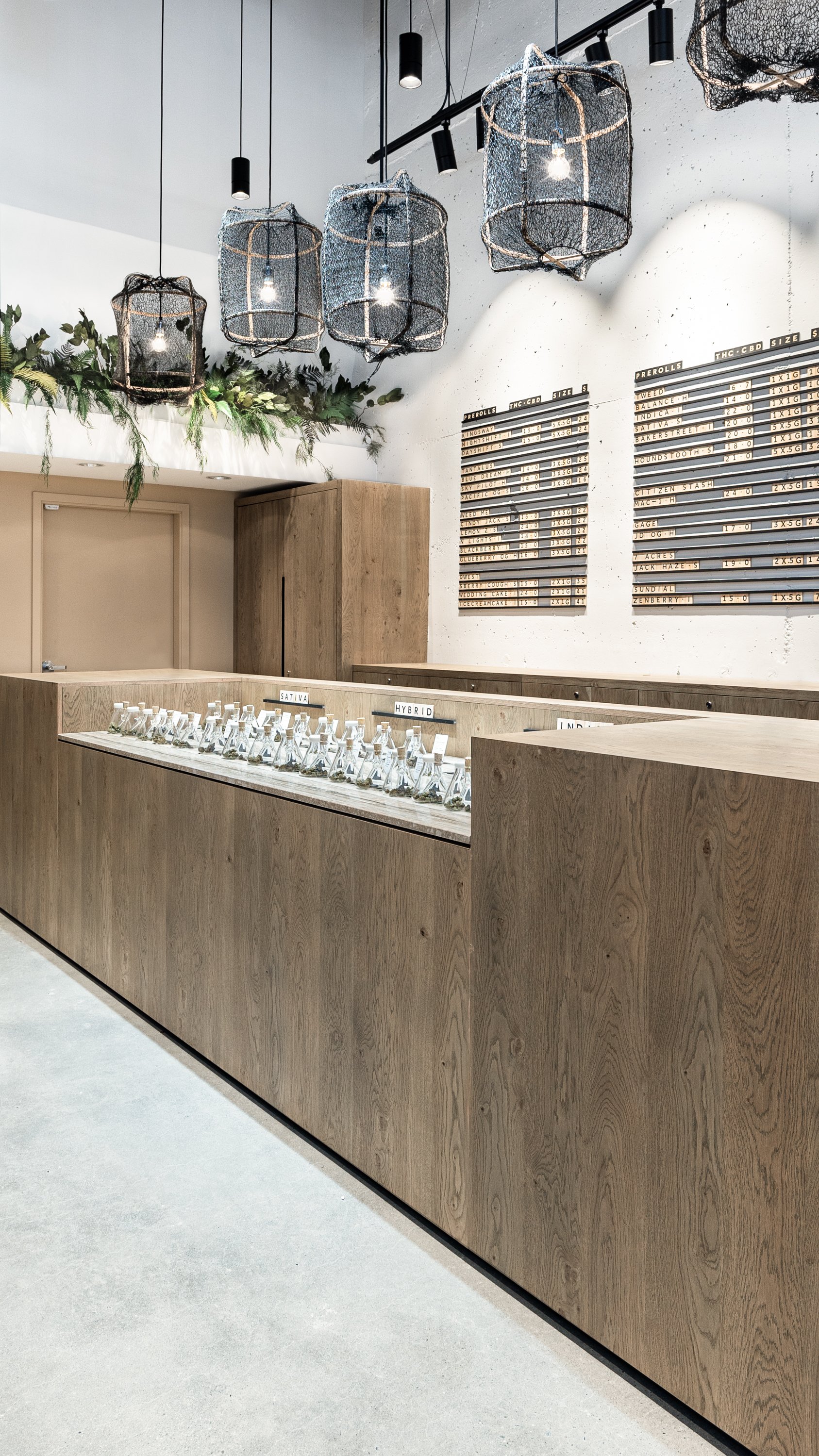 Interior of a modern dispensary with wooden counters displaying various cannabis products, black hanging pendant lights, a white wall with black and white boards with product information, and greenery decor overhead.