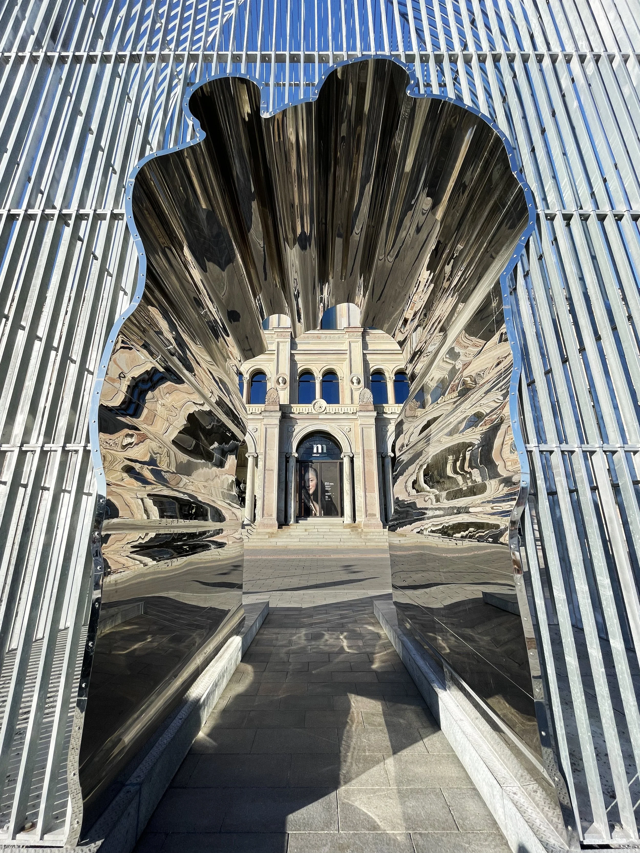 A metallic sculpture of a big shoe, creating a frame through which a historic building with arches and stairs is visible.