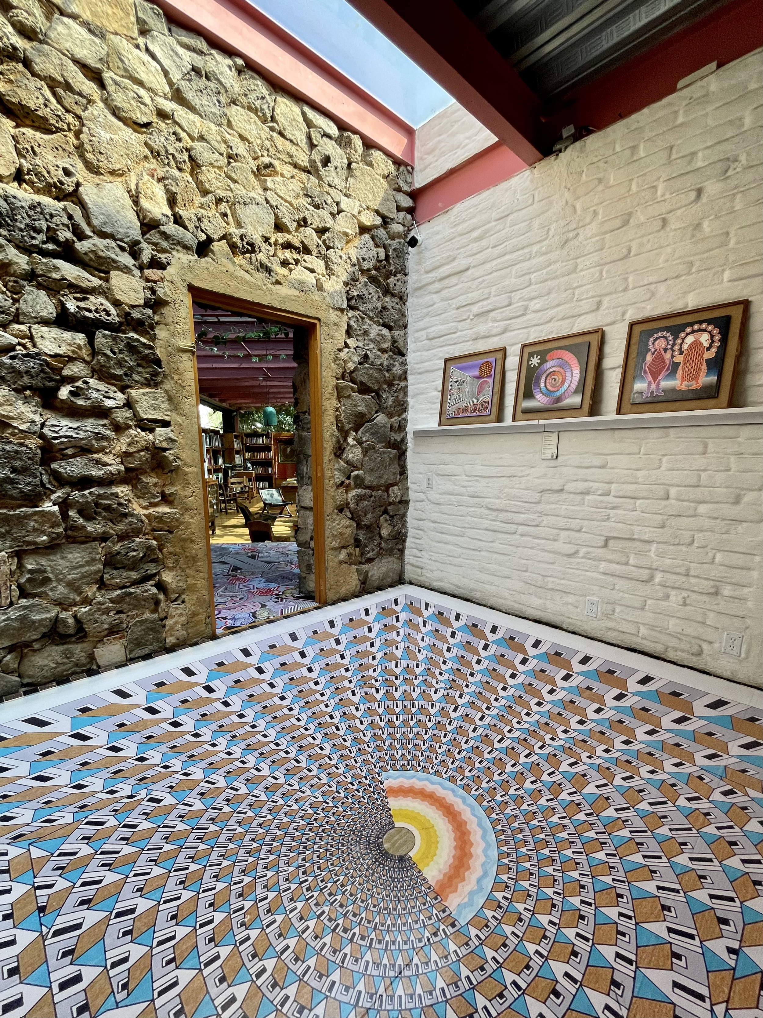 An interior room with a stone wall, white painted brick wall, and colorful geometric art print on the wall. The floor has a vibrant, optical illusion pattern with a spiral design in blue, brown, yellow, and black, creating a hypnotic effect.