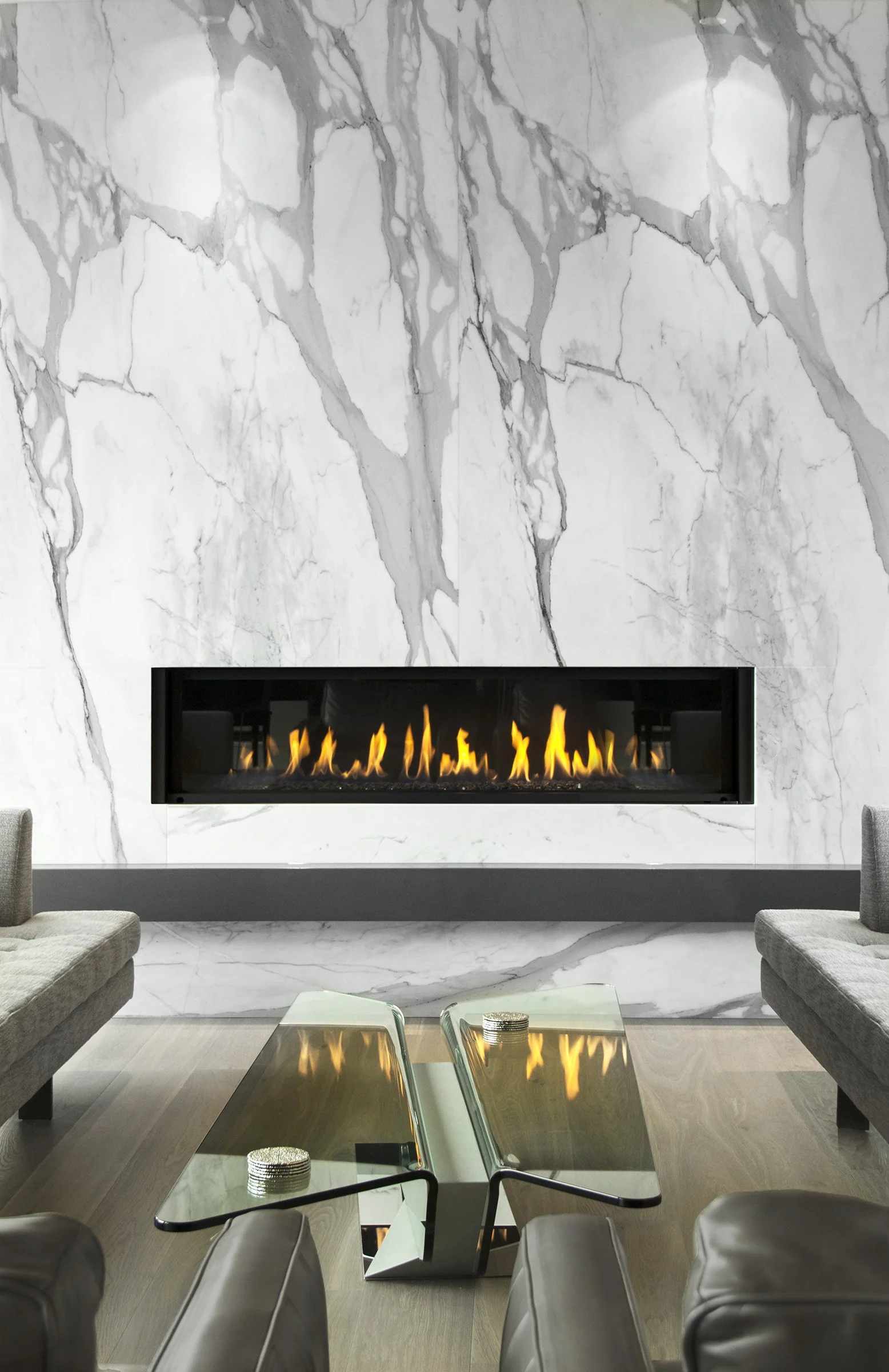Modern living room with a white marble wall featuring gray veining, a rectangular fireplace with flames, two gray sofas, and a glass coffee table with metallic accents.