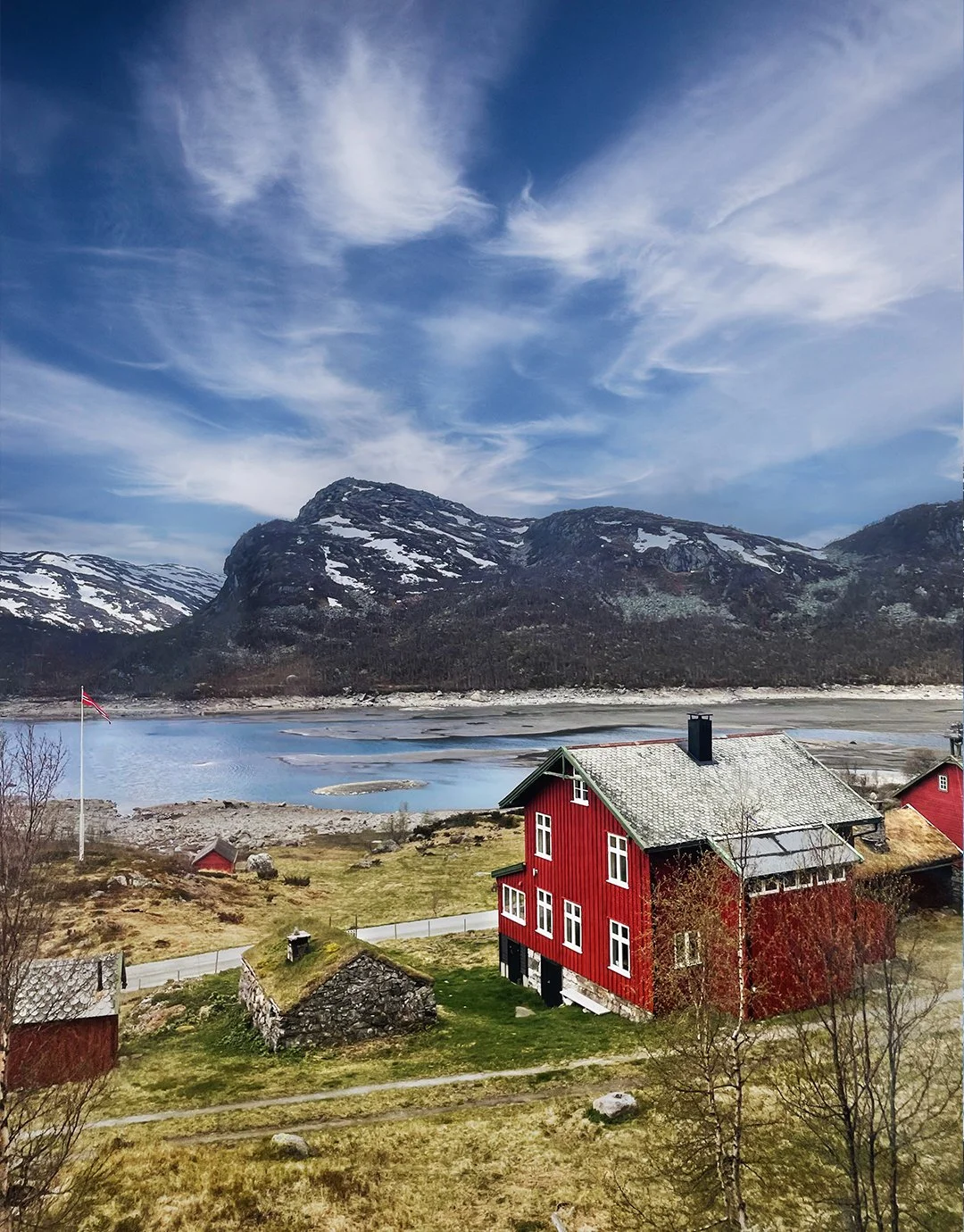 Scenic landscape of a red wooden house by a lake with snow-capped mountains and a partly cloudy blue sky in the background.