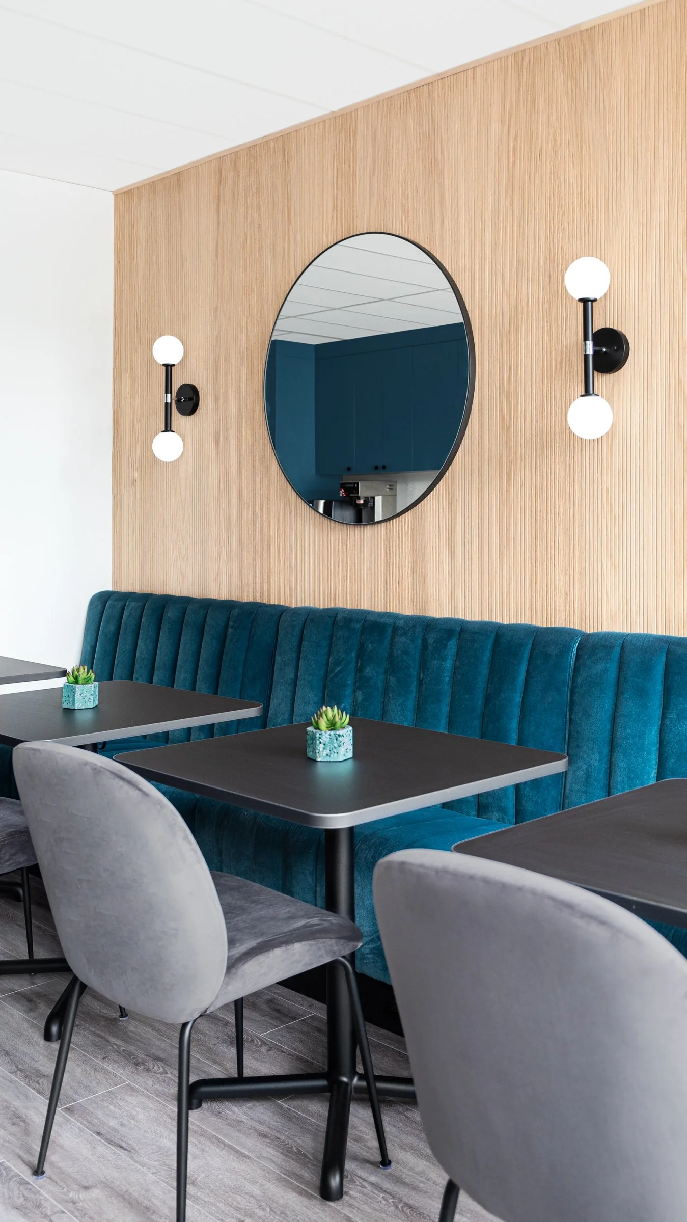 Modern interior of a restaurant with blue velvet booth seating, grey chairs, black tables, decorative plants, a round mirror, and black wall-mounted light fixtures.