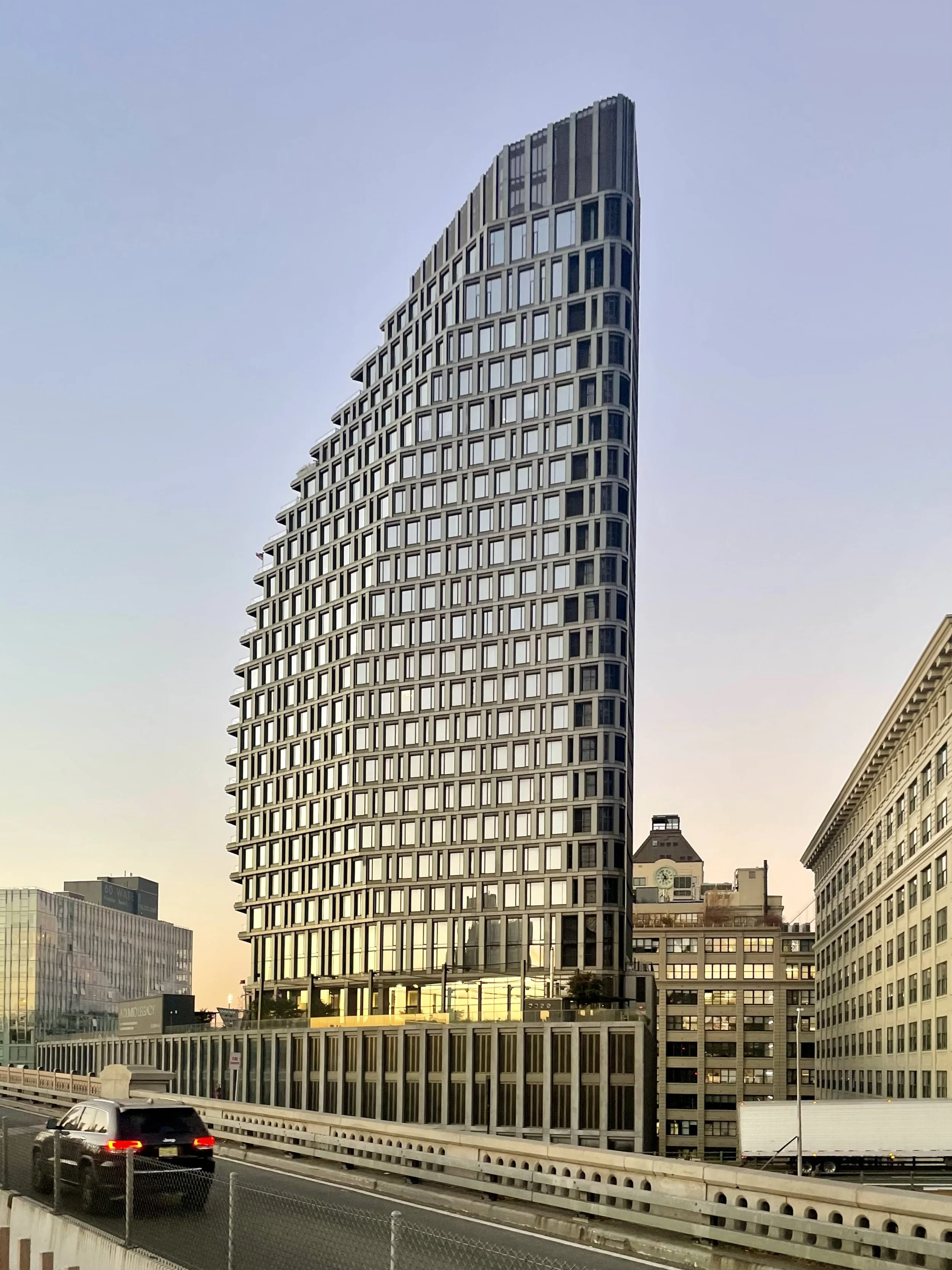 Tall, modern high-rise building with unique, curving architectural design on a city street during sunset.