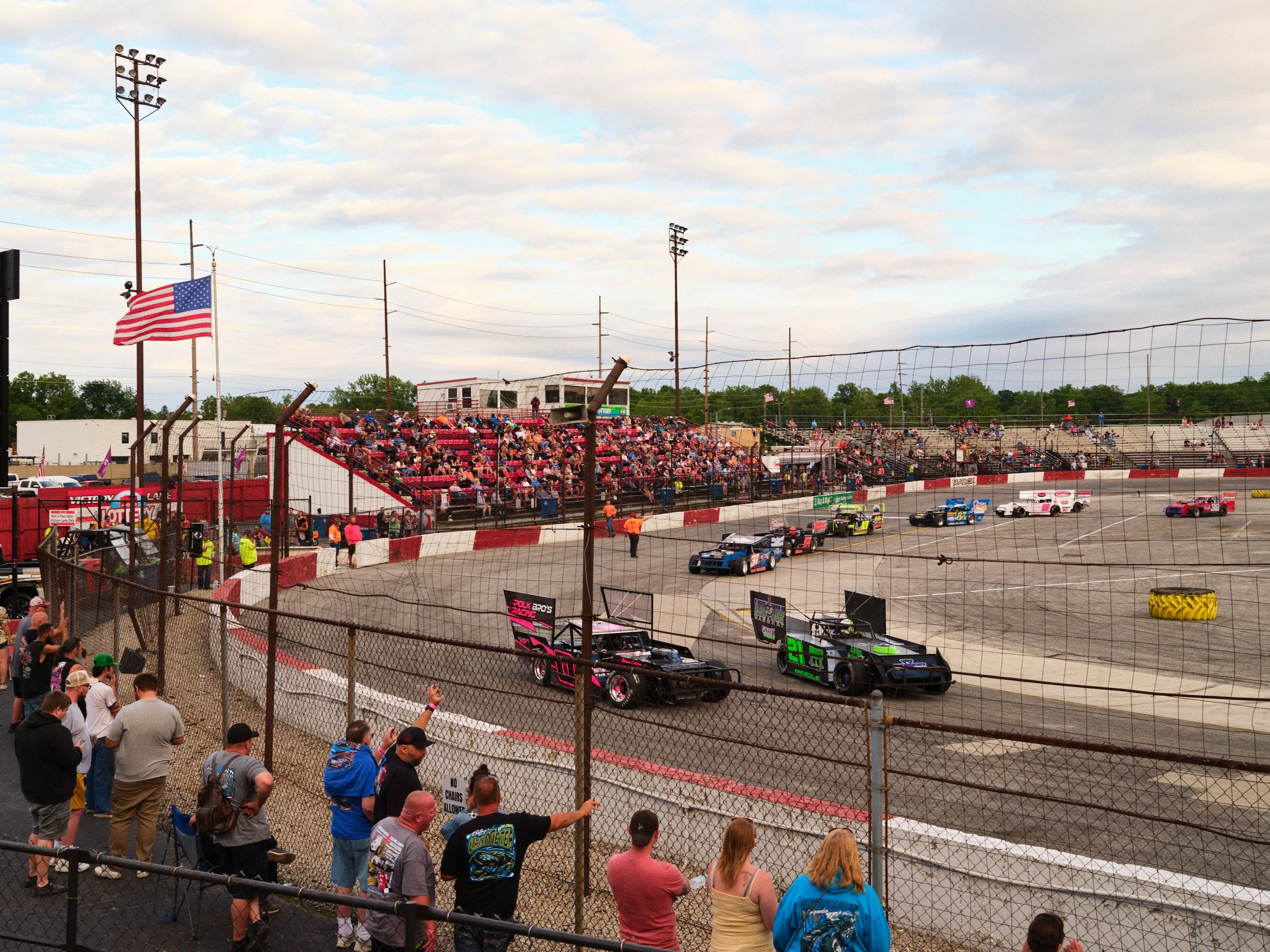 A dirt track race car event with cars racing on the track, surrounded by a chain-link fence, with spectators in the stands and on the ground. The American flag is flying on a flagpole near the track.