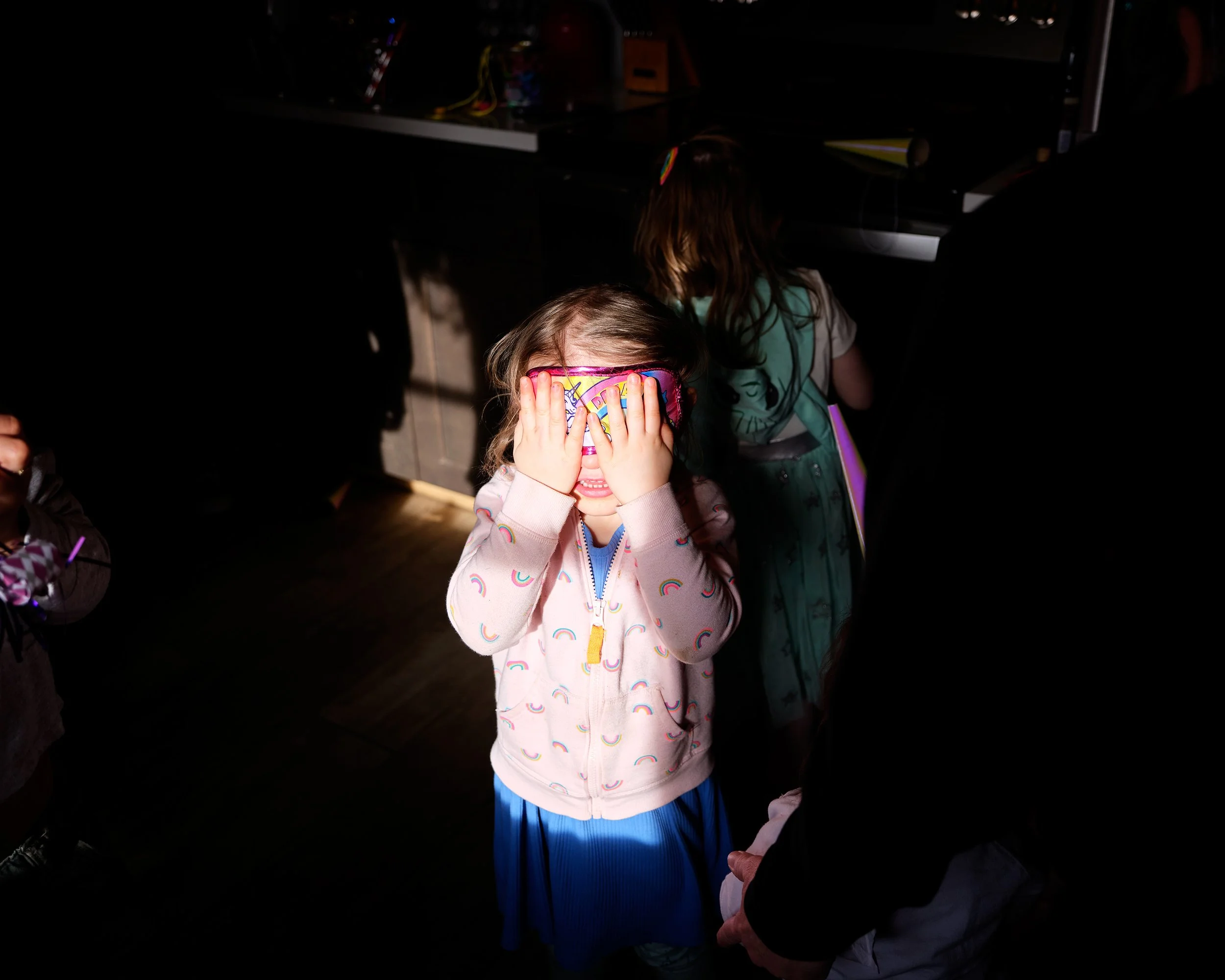 A young girl with long hair, wearing a rainbow-colored eye mask and a pastel jacket with rainbow patterns, covering her face with her hands in a dimly lit room with sunlight streaming in.