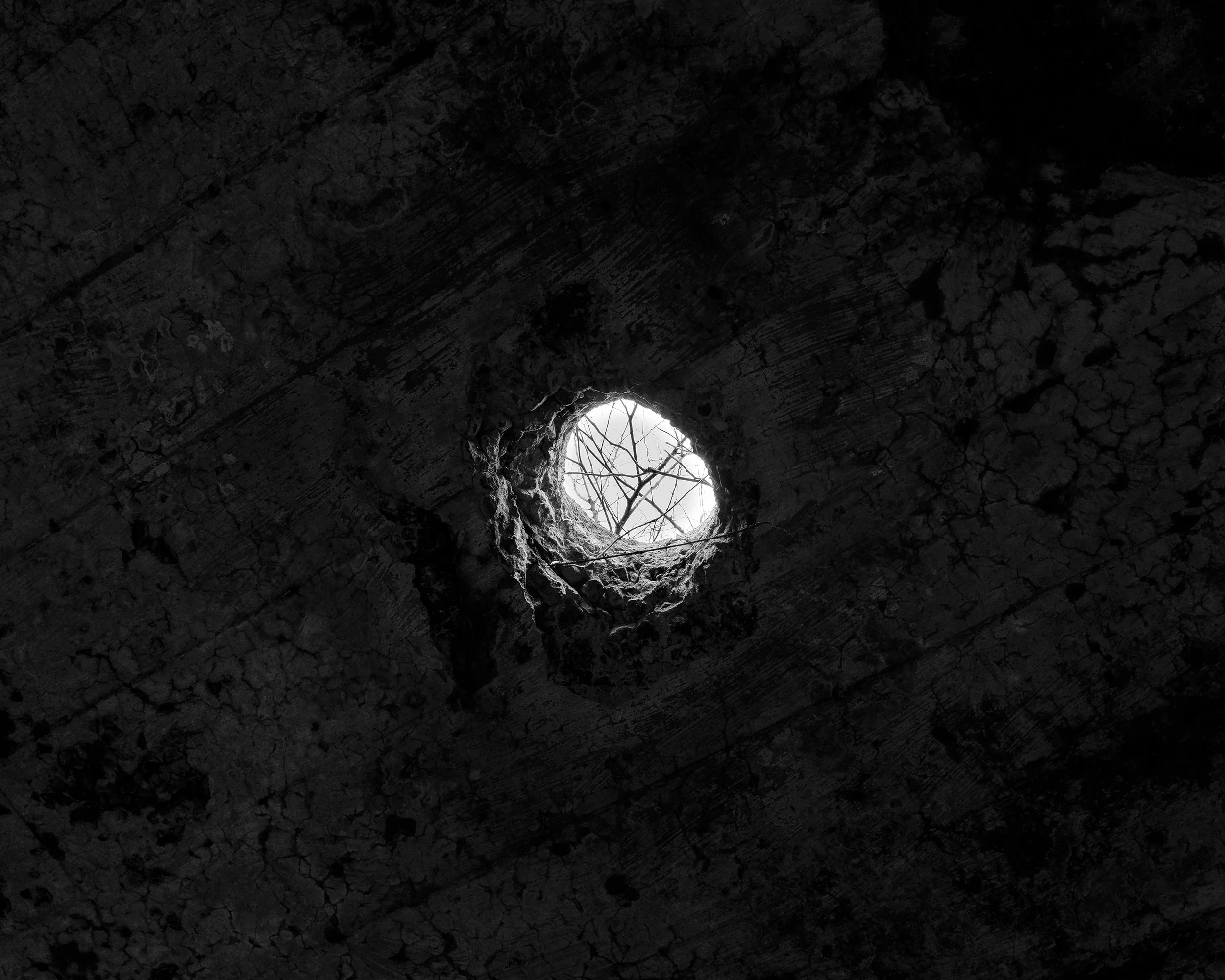 Looking up through a circular hole in a dark, weathered surface, with some branches visible through the opening.