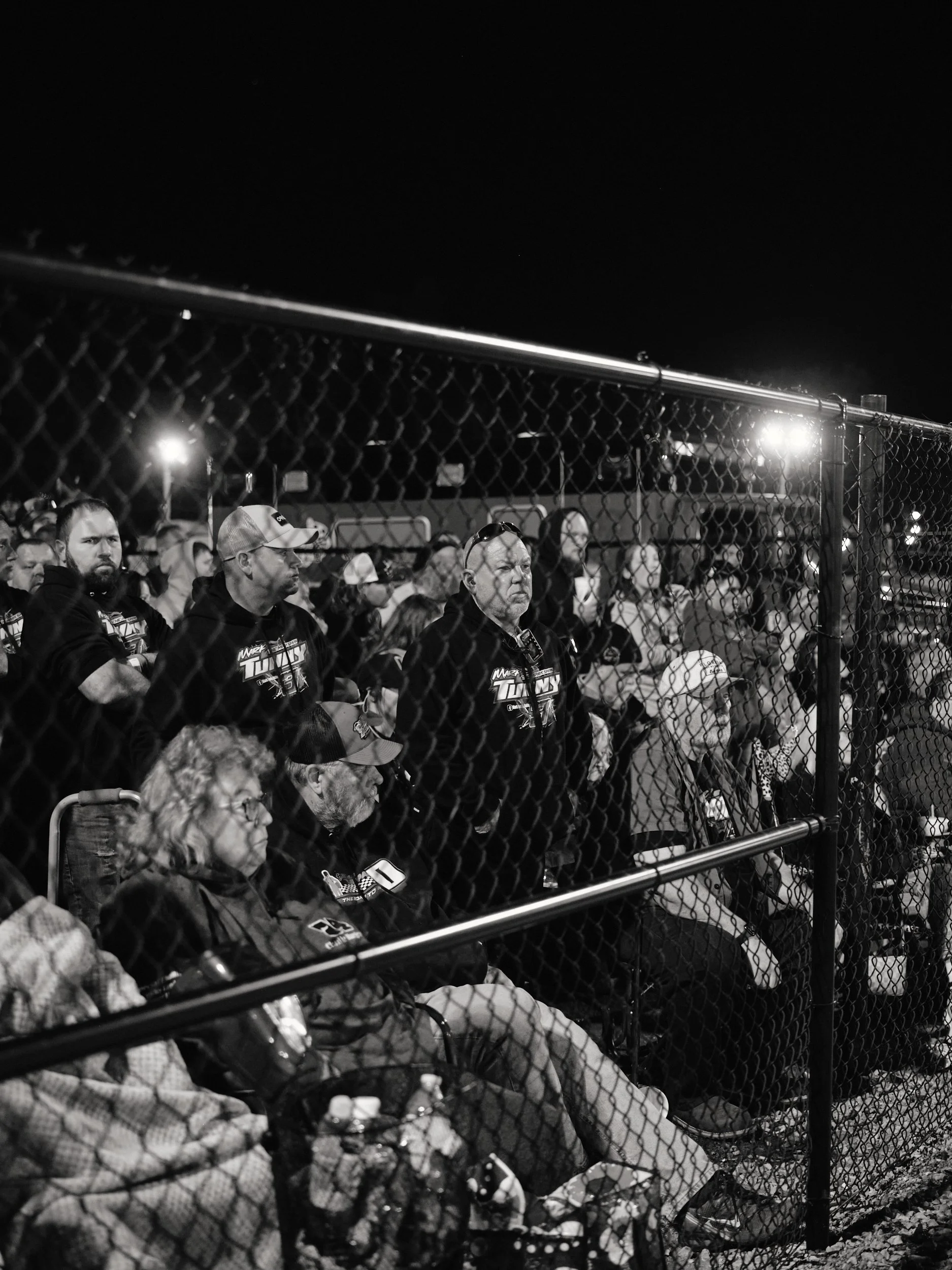 Nighttime outdoor sports event with spectators behind a chain-link fence, illuminated by bright stadium lights, in black and white.