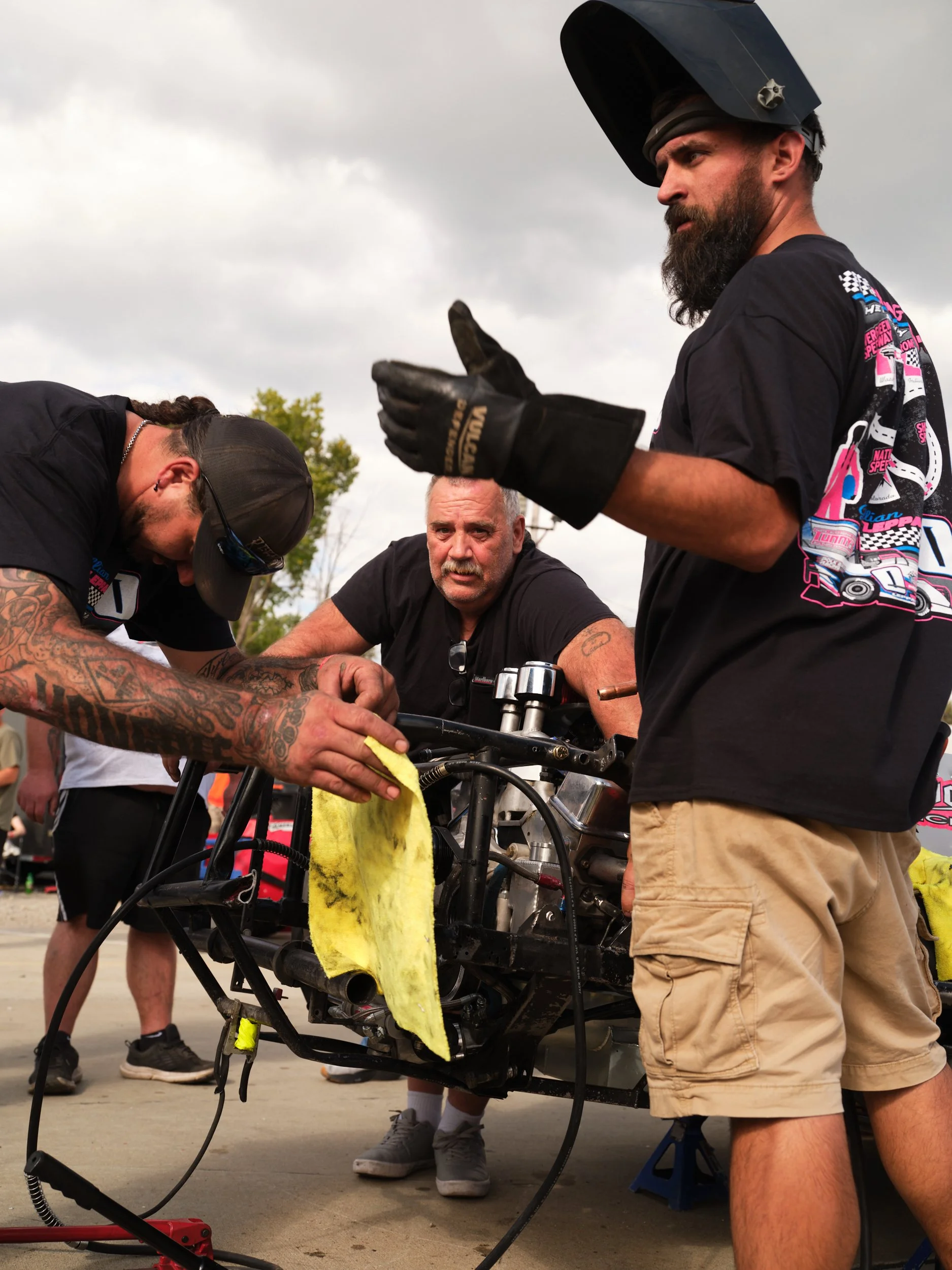 Three men are working on a motorcycle engine outdoors, with one of them holding a yellow cloth and the other two observing. They are dressed casually, and the sky is overcast.