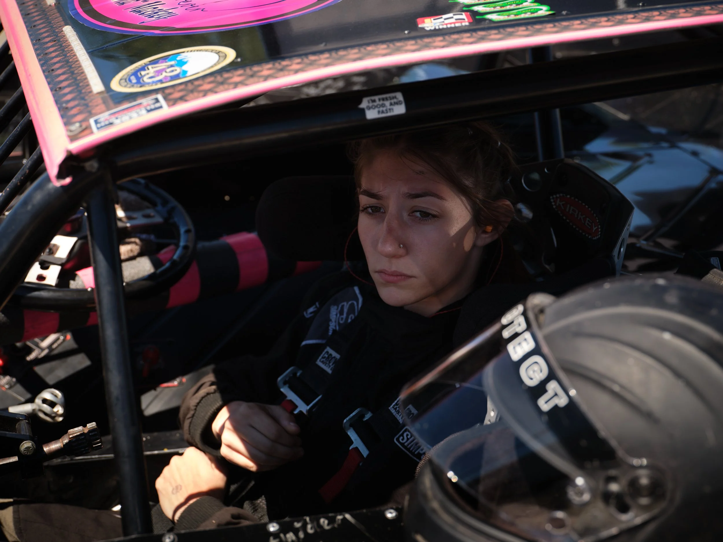 A young woman sitting inside a race car, wearing a black race suit with sponsor patches, and holding her hands near her chest. She has a focused expression and is positioned behind a clear helmet visor.