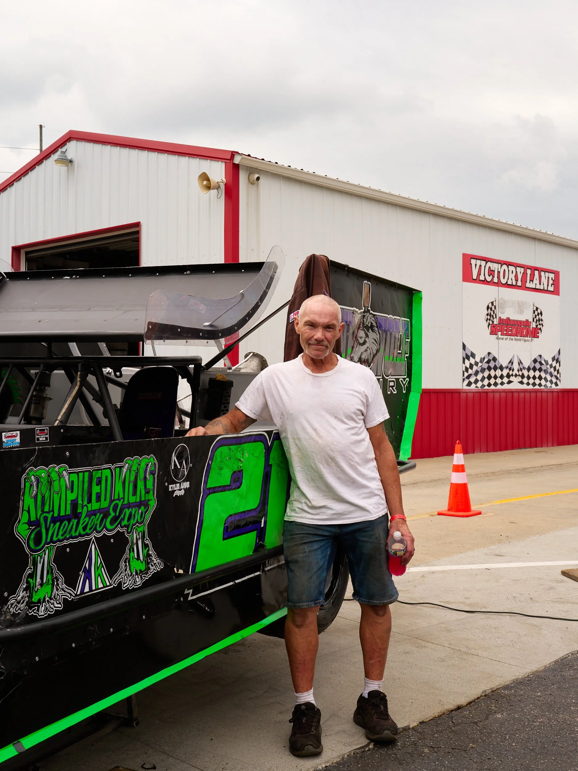 Older man in white t-shirt and shorts standing next to a race car at a speedway, with the track and a building labeled 'Victory Lane' in the background.
