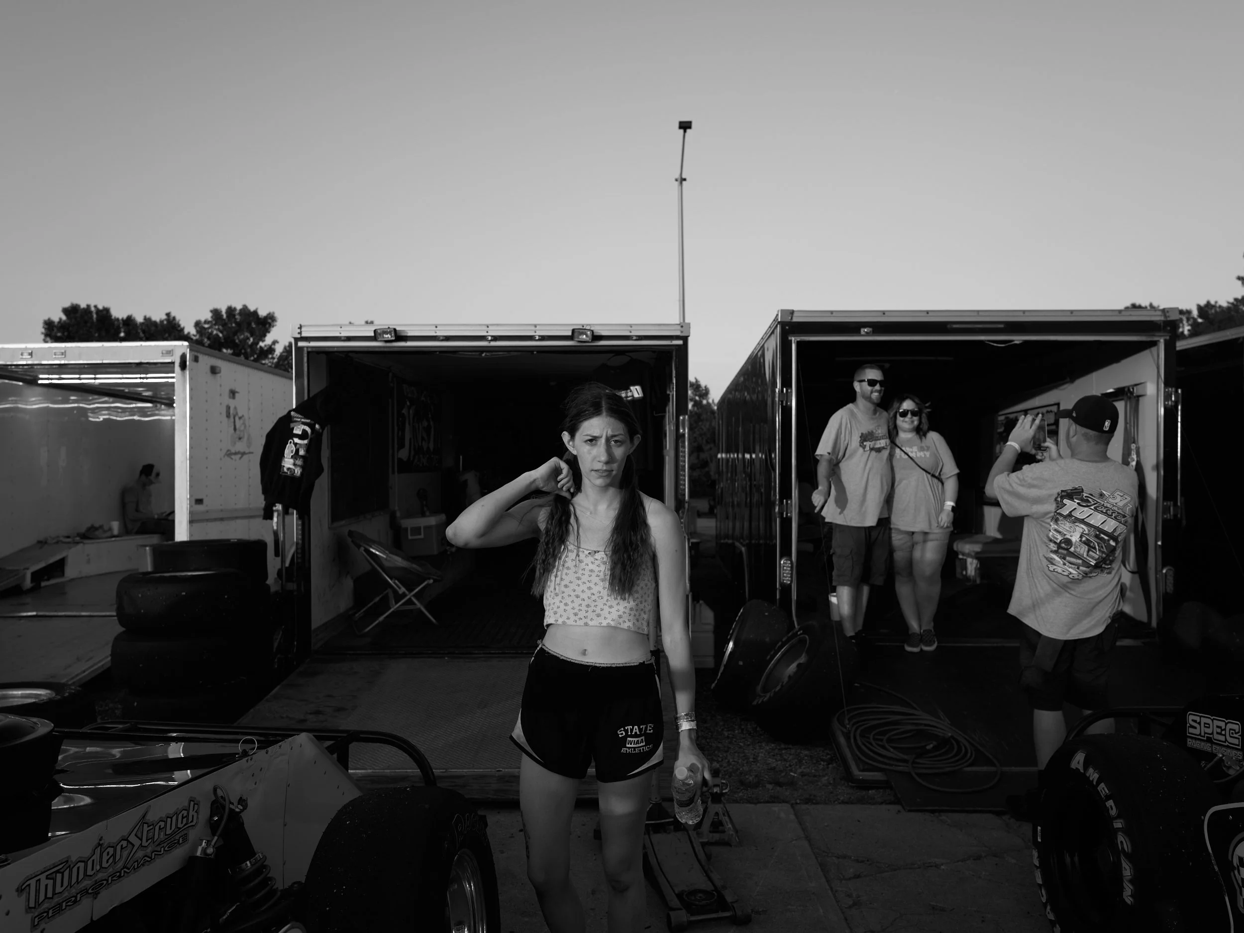 People in a racing paddock area with open trailer trucks, a woman in the foreground holding a water bottle and looking at the camera, and two others near the trucks.