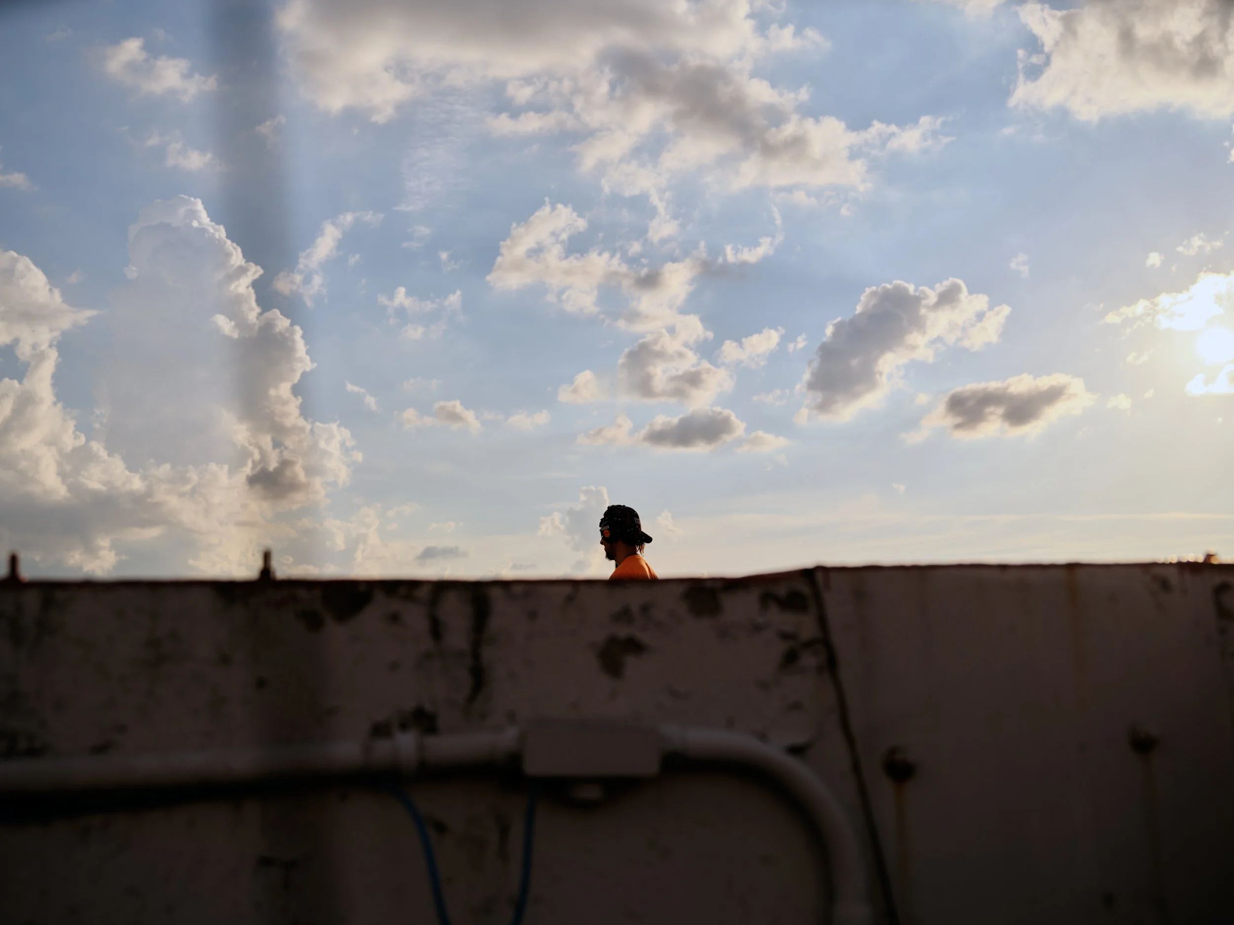 Side profile of a person with dark hair wearing an orange shirt and sunglasses, standing outdoors near a rooftop with a cloudy sky background and the sun shining.