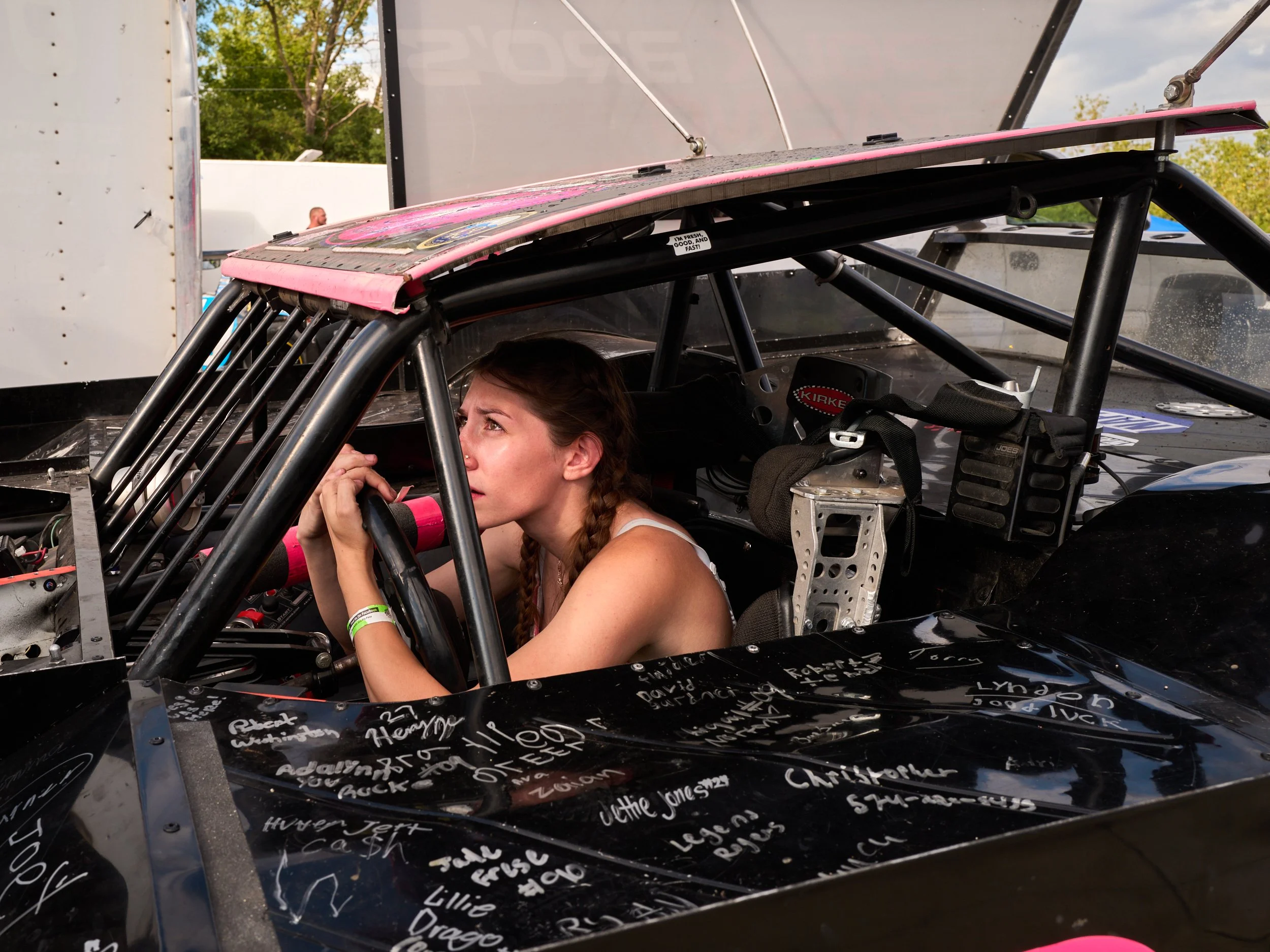 A woman inside a race car, holding the steering wheel and looking out, with handwritten names and messages on the black dashboard.