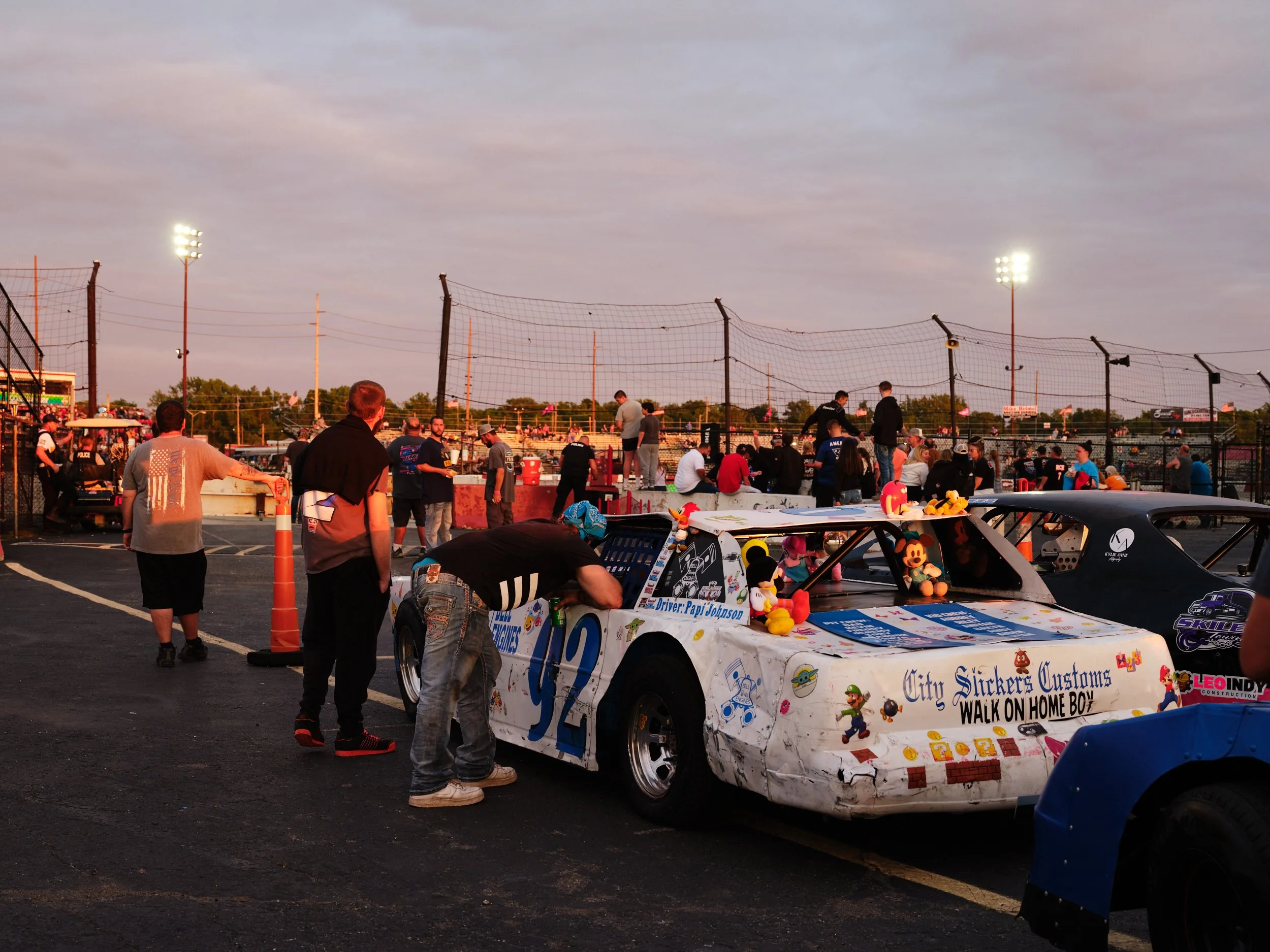 A group of people around a race car decorated with plush toys and stickers at a race track during sunset.