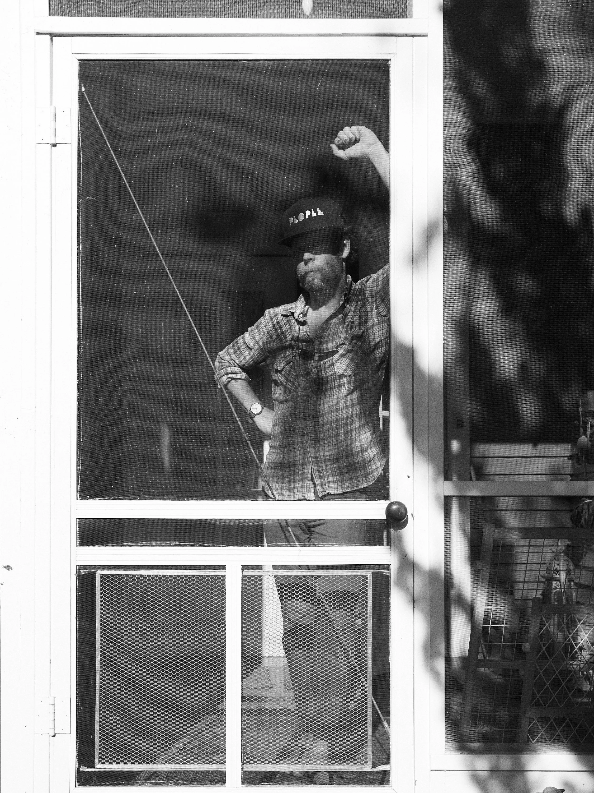 A person standing behind a screen door, wearing sunglasses, a plaid shirt, a watch, and a hat that says 'FOLK.'