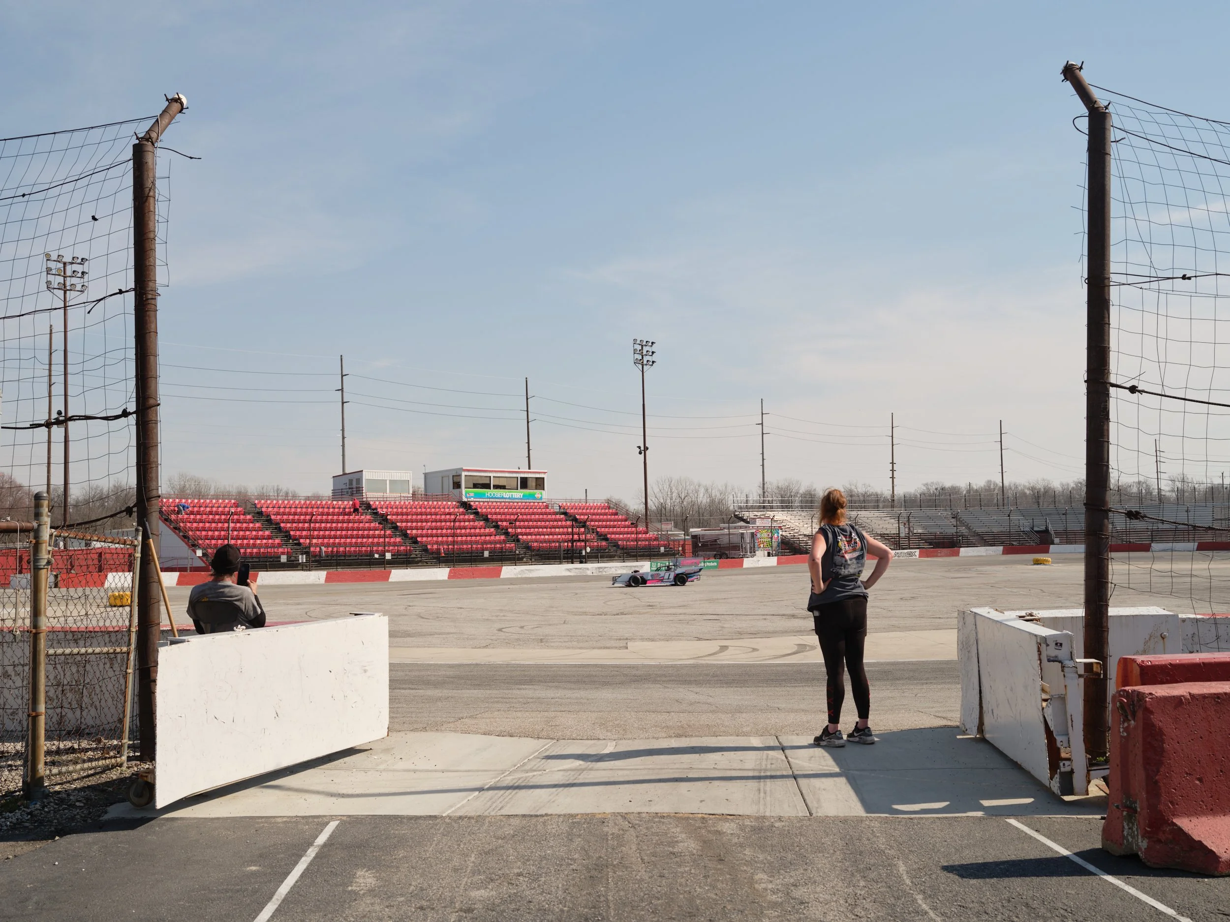Two people at a race track, one taking a photo and the other standing with hands on hips, with a race car speeding on the track in the background.