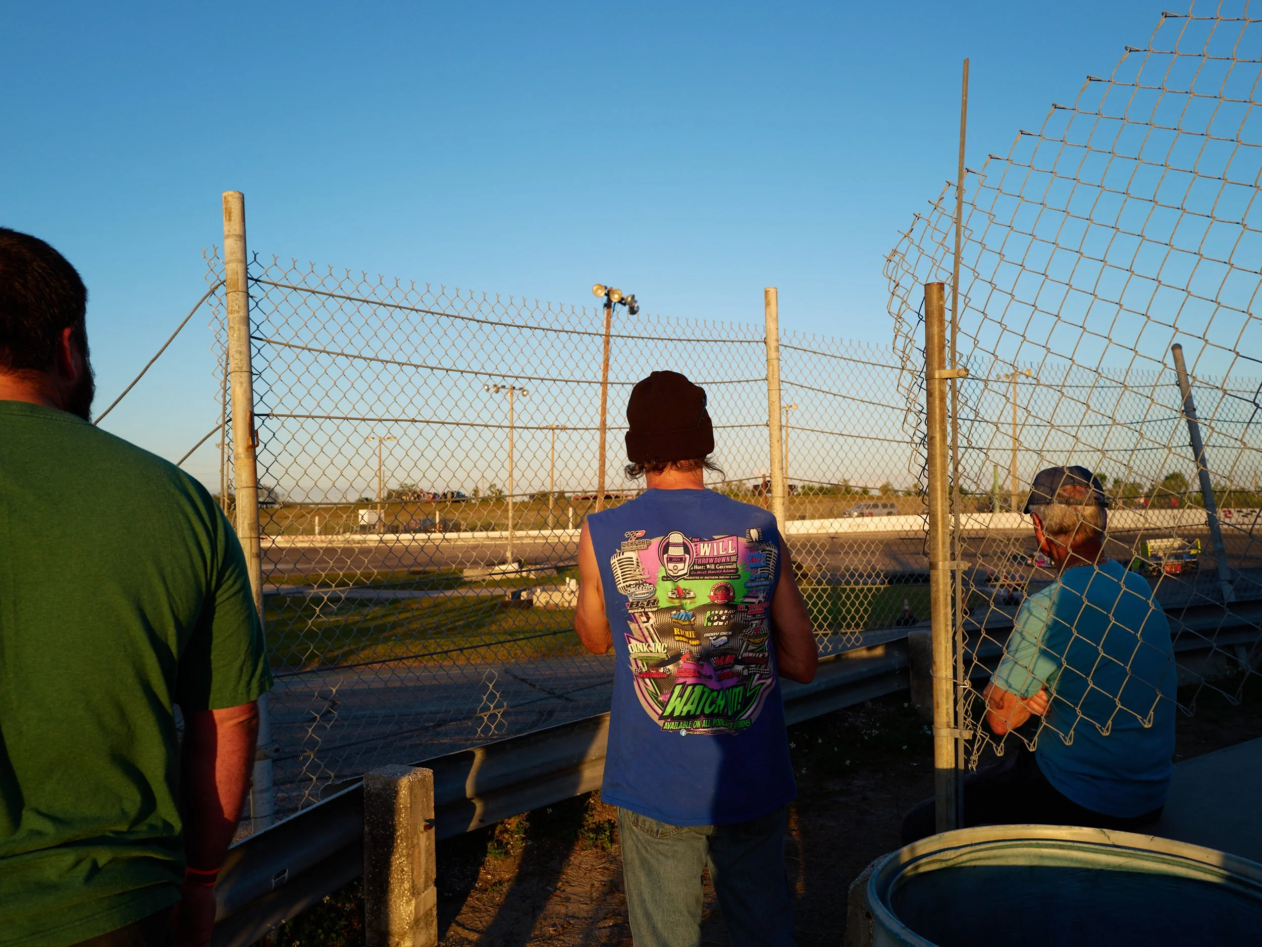 Three men stand behind a chain-link fence at a race track, watching cars on the track during sunset.