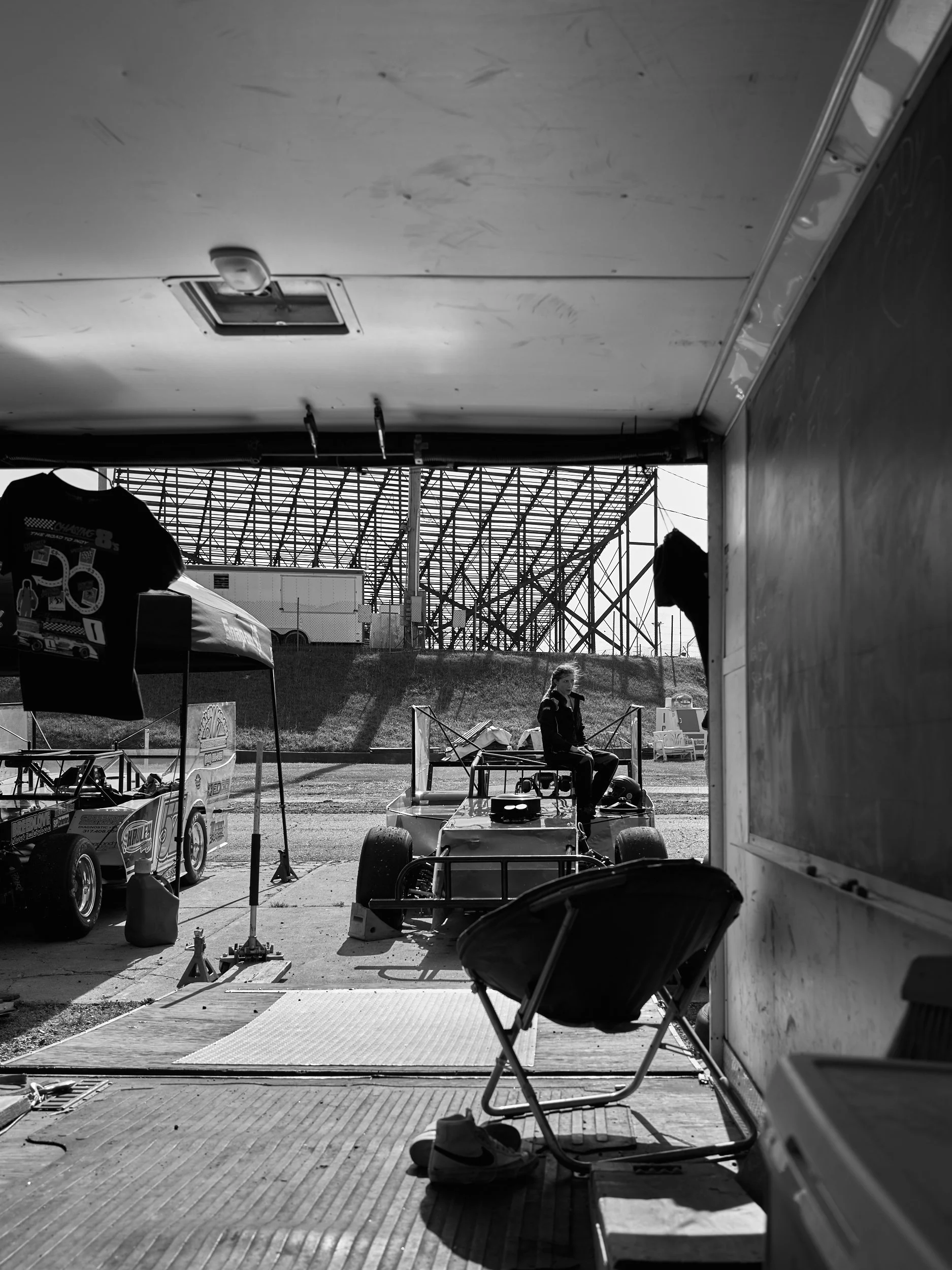 Black and white photo of a person sitting on a small race car under a garage, with a racetrack and grandstand in the background.