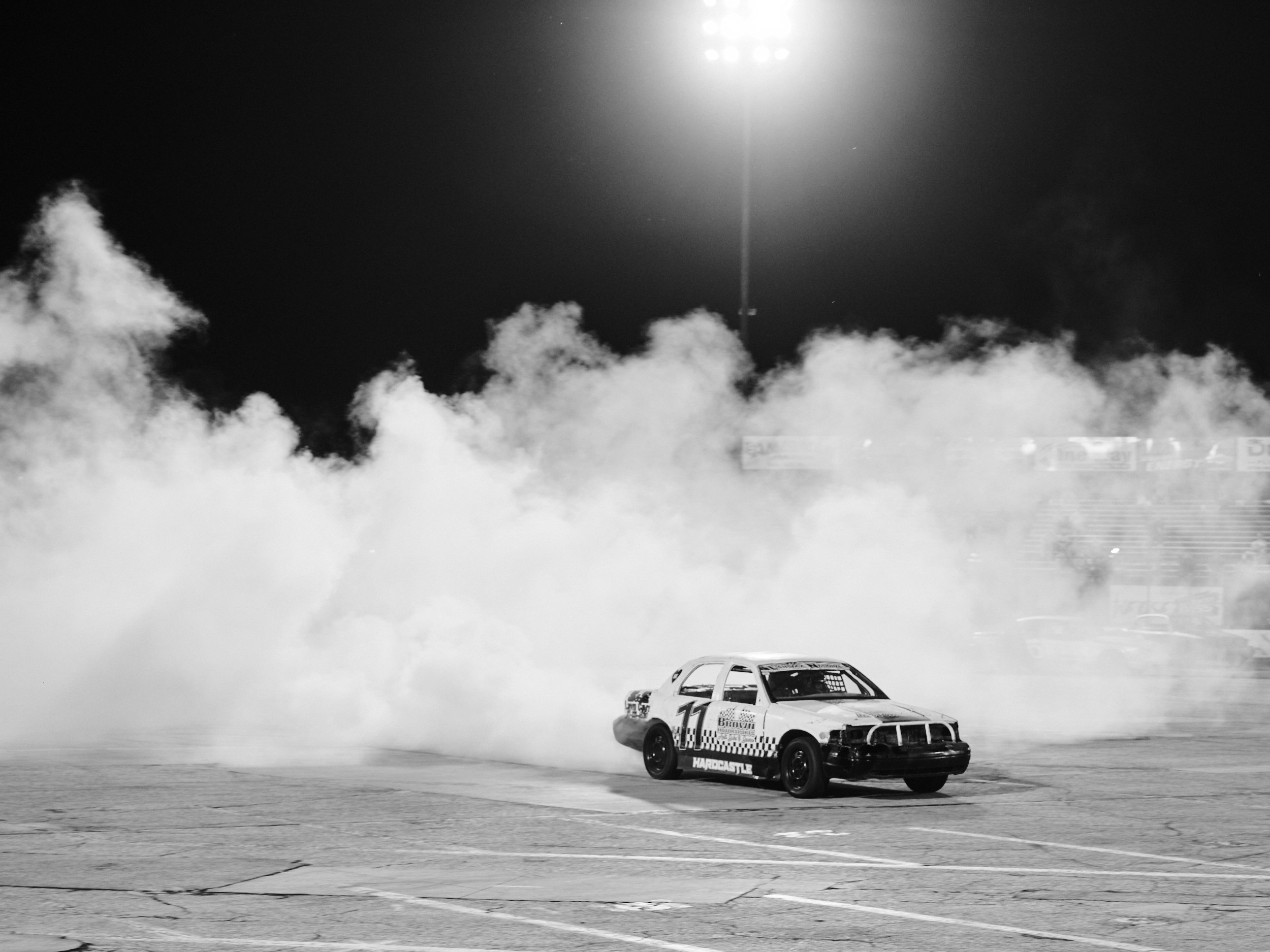 A race car performing a burnout at night, creating a large cloud of smoke in an empty parking lot under excess lighting.