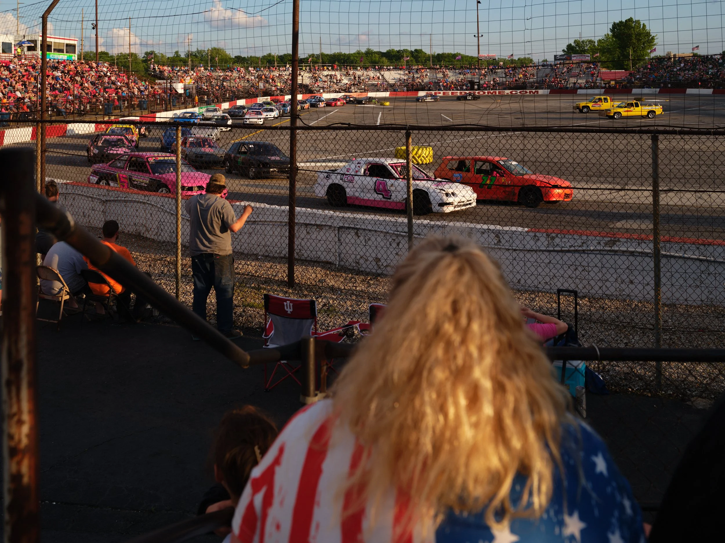 People seated in front of a racetrack observing race cars on the track with spectators in the stands.