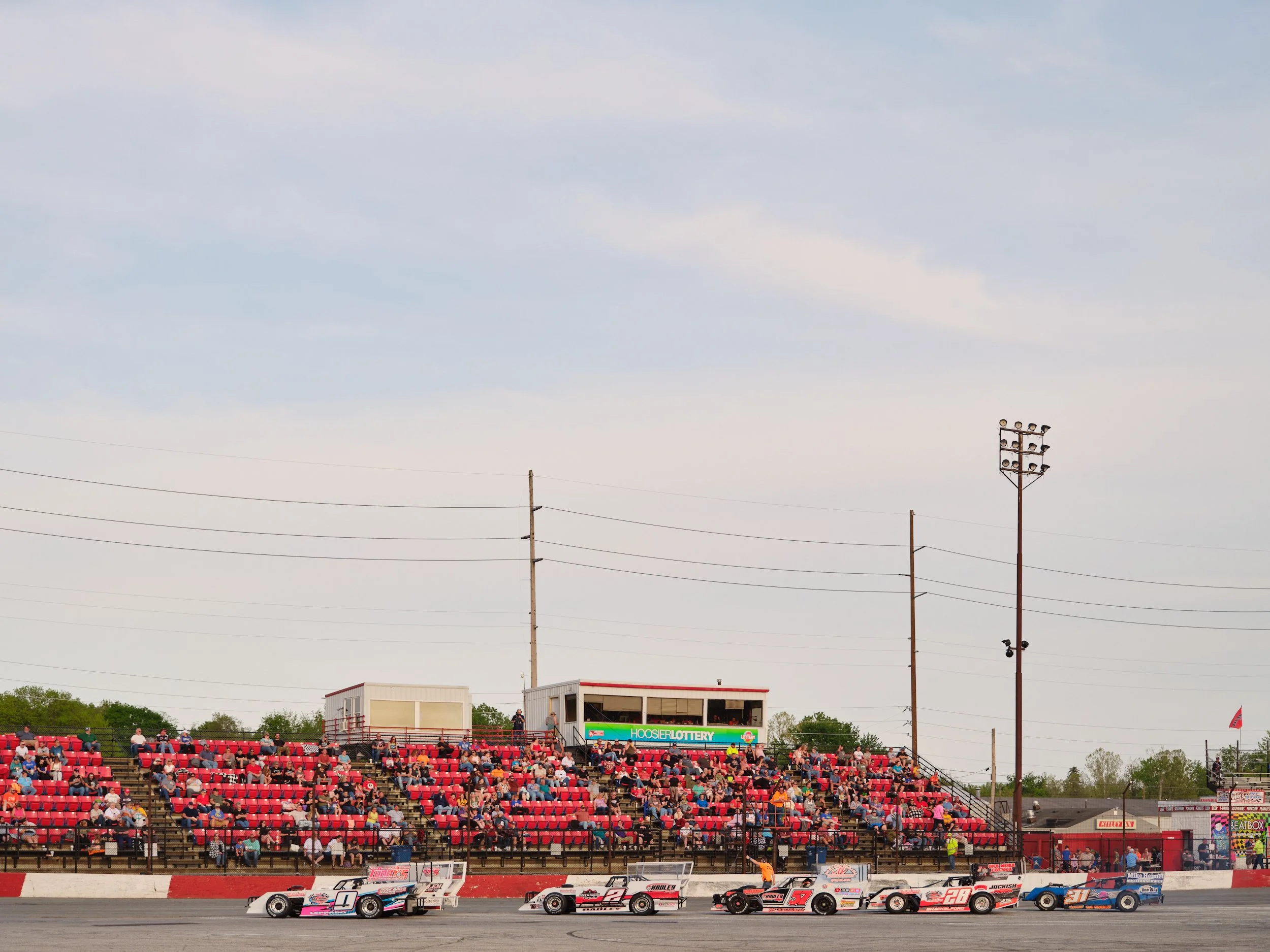 Race cars on a track with grandstand full of spectators and lights, under a partly cloudy sky.