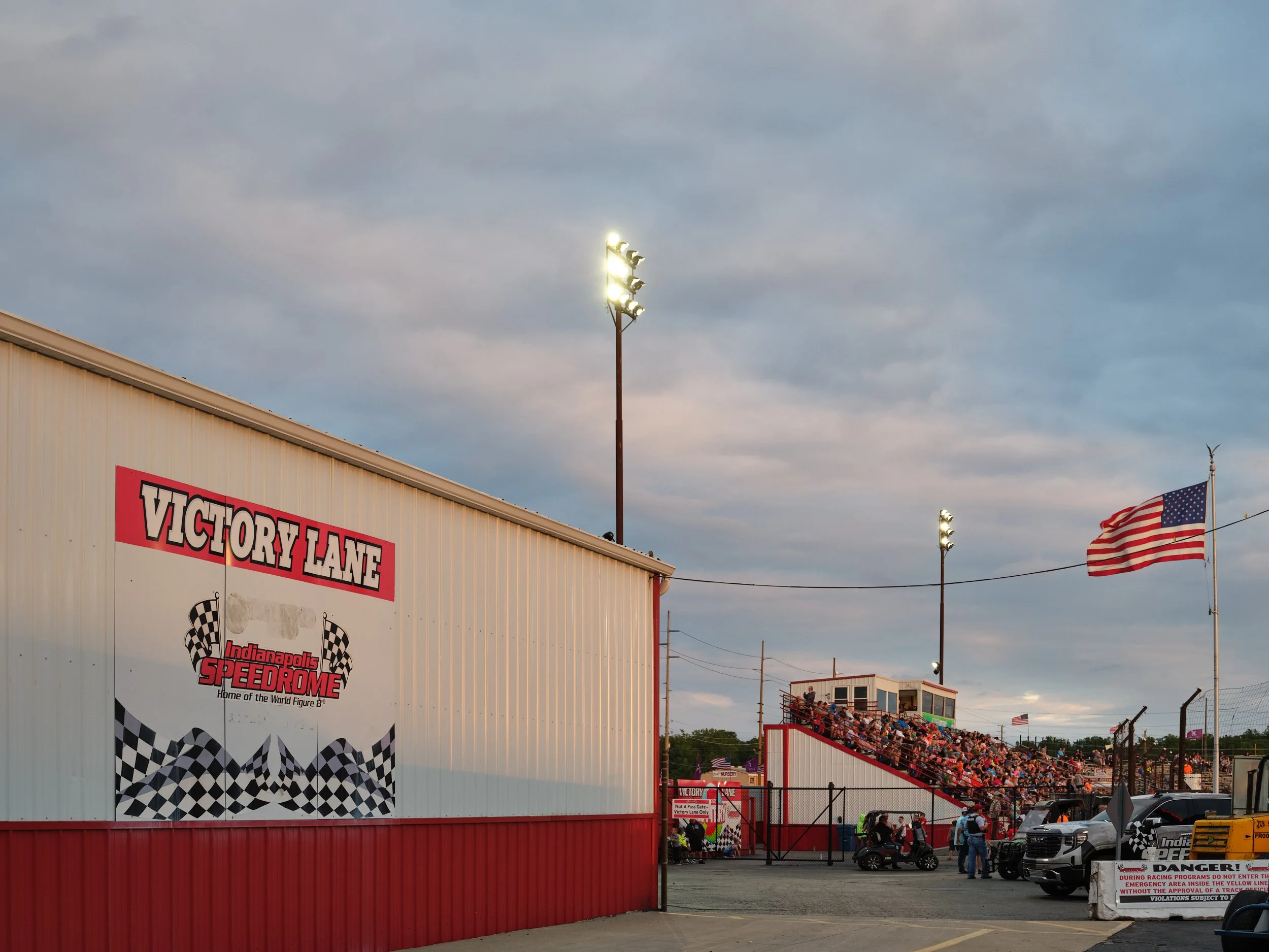 The Indianapolis Speedway Victory Lane with grandstands filled with spectators, American flags flying, and bright floodlights illuminating the area under a partly cloudy sky.