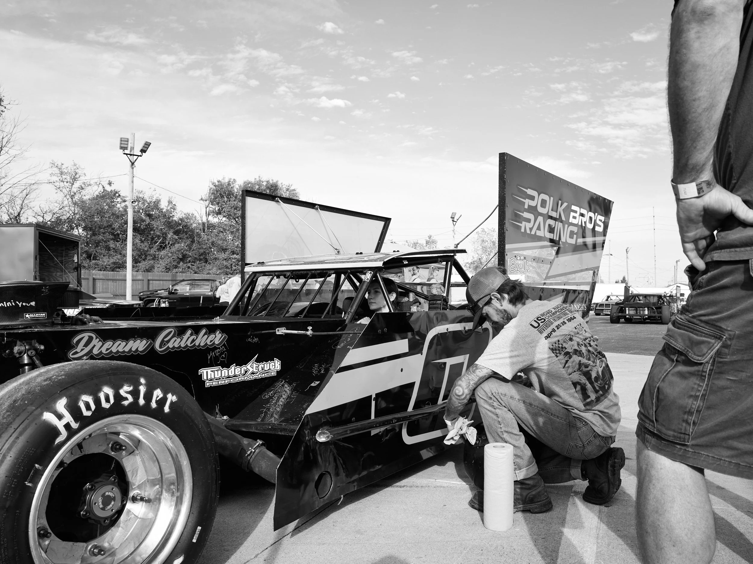 A man with tattoos and a cap kneeling next to a race car, adjusting or working on it, in a parking lot or area with other race cars and vehicles in the background.