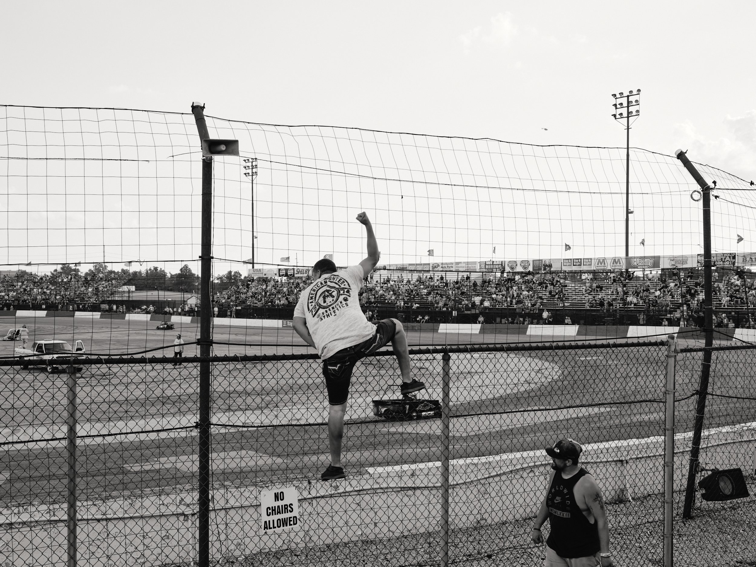 A man climbing a chain-link fence at a racetrack while raising his right fist in the air.
