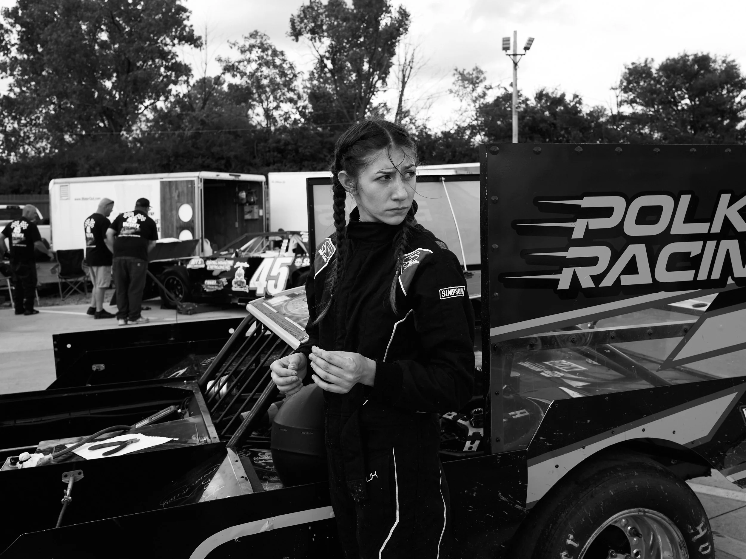 Black and white photo of a woman in racing gear standing beside race cars and team members in the background at a racing event.