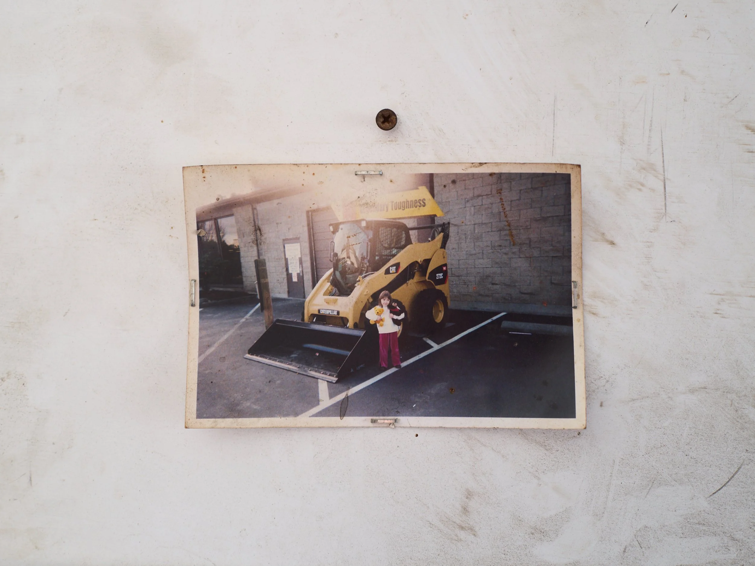 Photograph of a young girl holding a stuffed animal, standing in front of a construction vehicle parked outside a building.