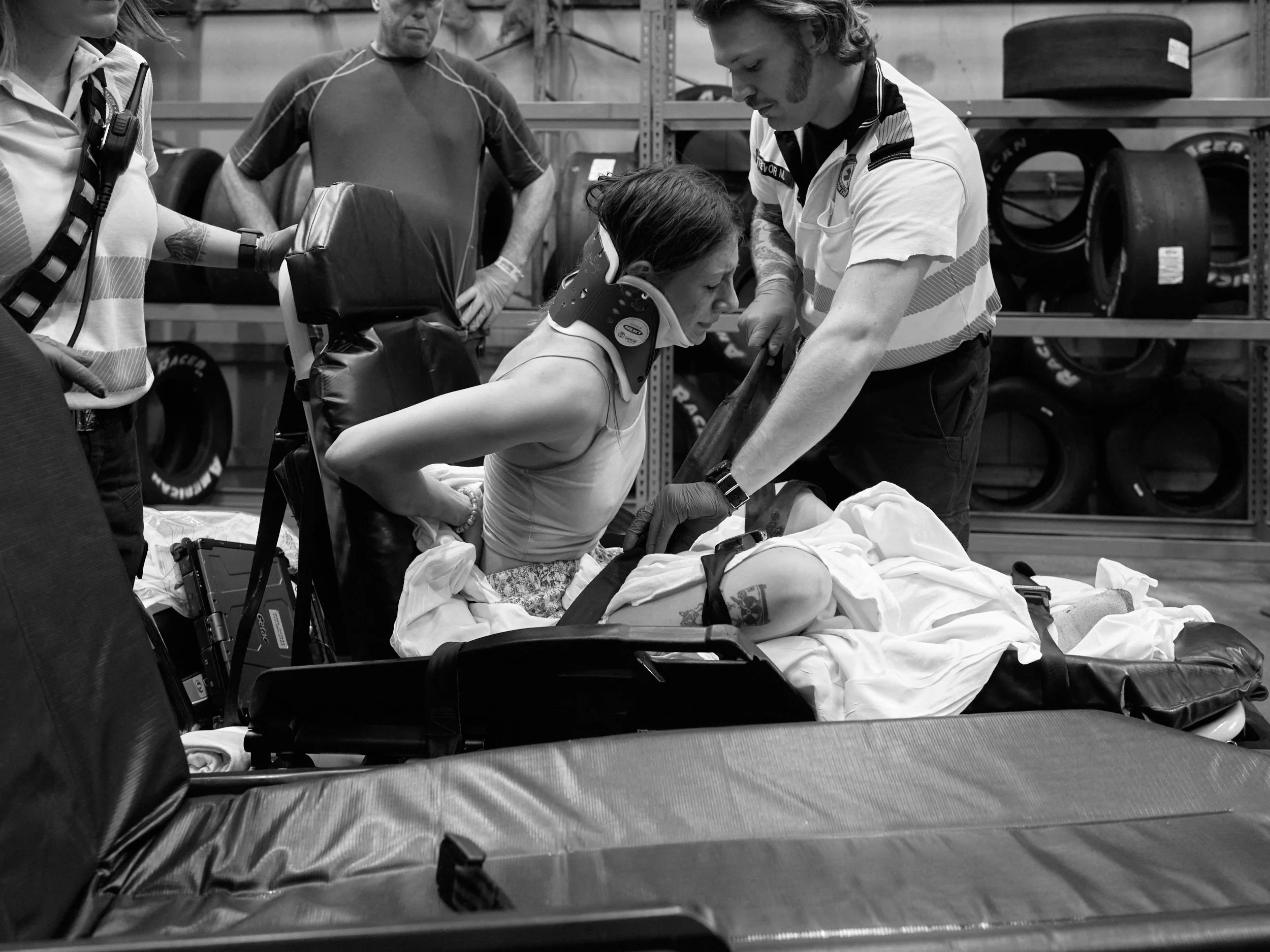 A female race car driver with a neck collar and arm tattoo is receiving medical attention from emergency personnel in a racing garage, with tires stored on shelves in the background.