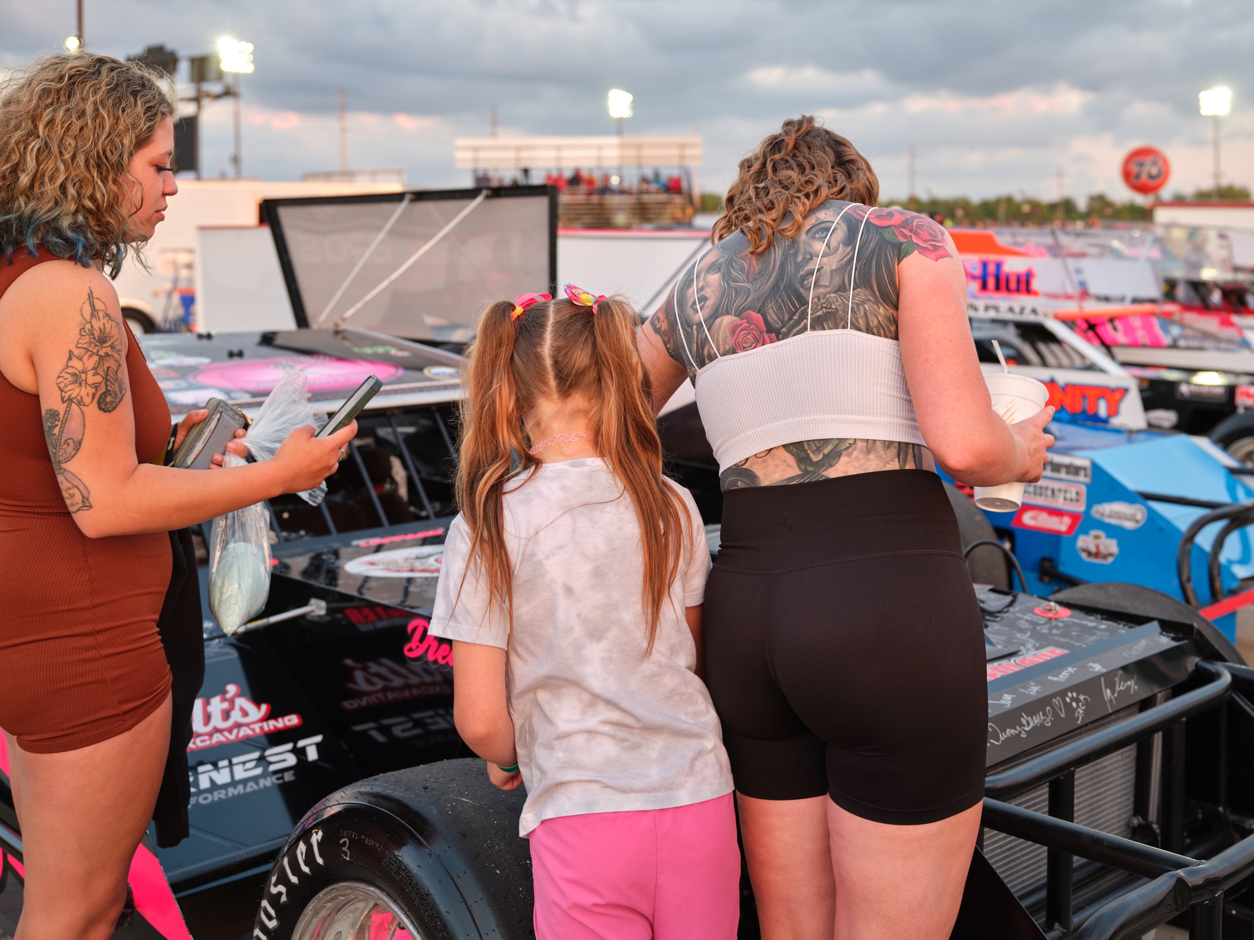 Three women and a girl standing by race cars at a racing event, with the women looking at their phones or holding drinks.