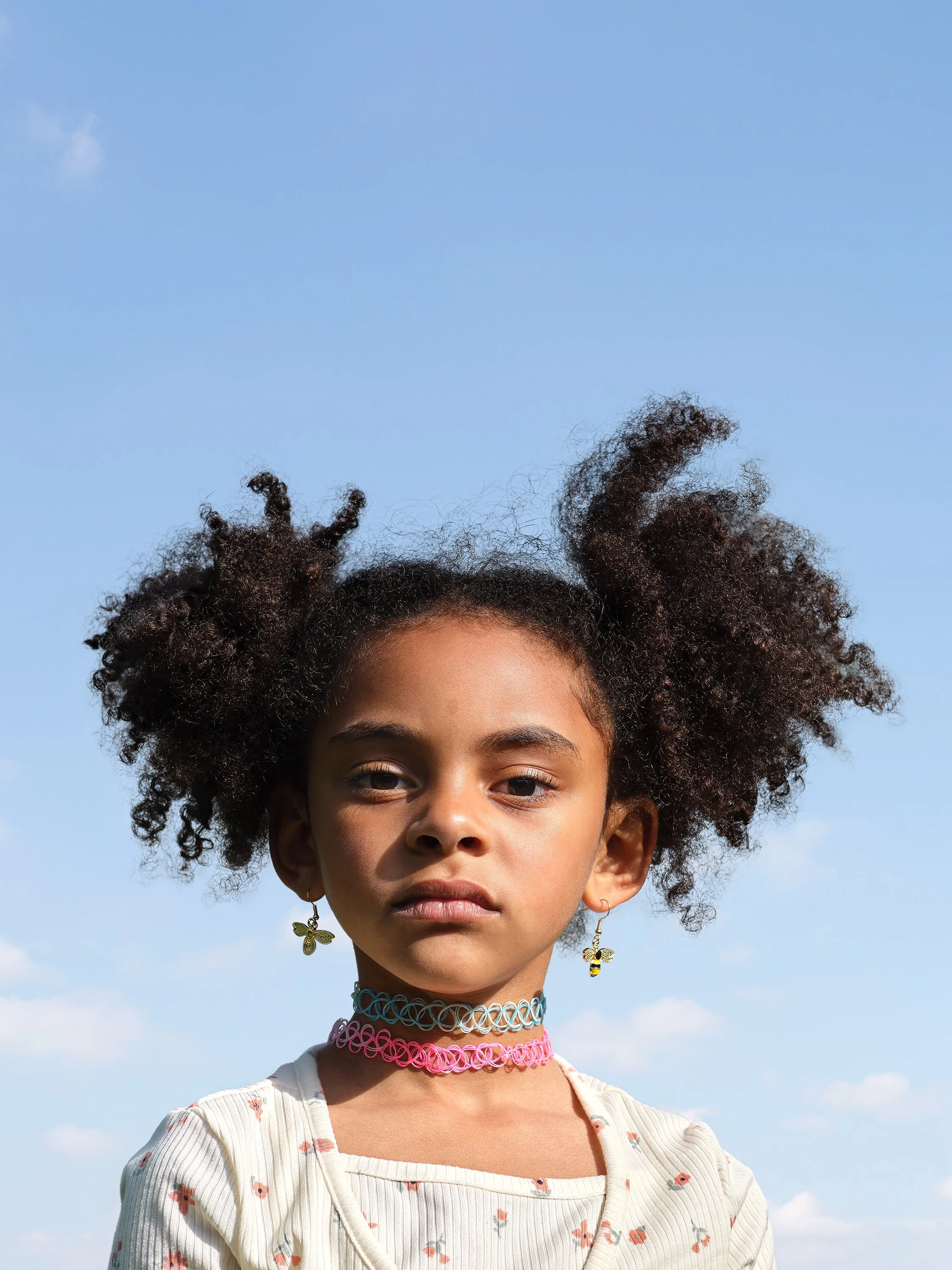 A young girl with curly hair styled in puffs, wearing earrings, a colorful choker, and a patterned shirt, stands outdoors against a blue sky with clouds.