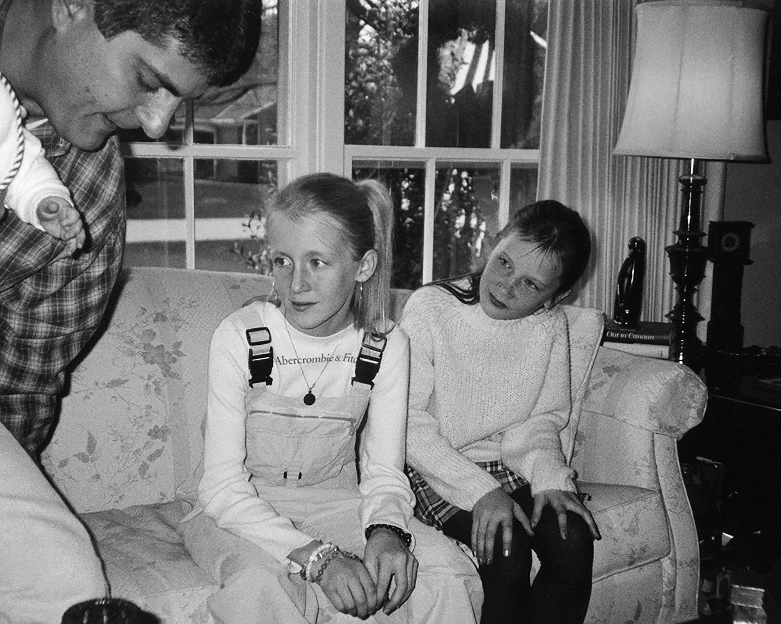 Two young girls sitting on a floral-patterned sofa, with a man standing nearby, in a room with a large window and a lamp in the background.