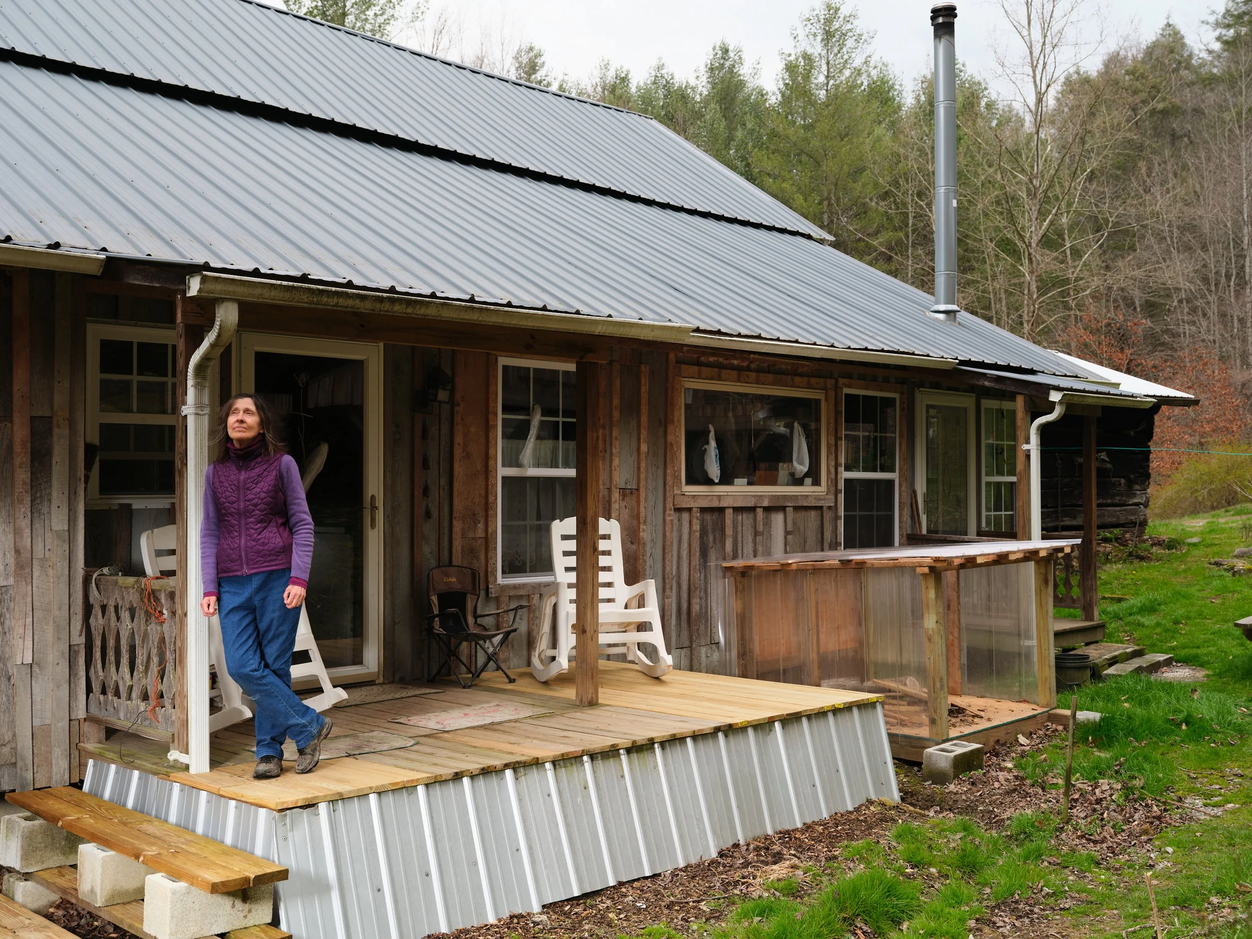 A woman standing on a wooden porch of a rustic house with a metal roof, surrounded by trees and greenery.