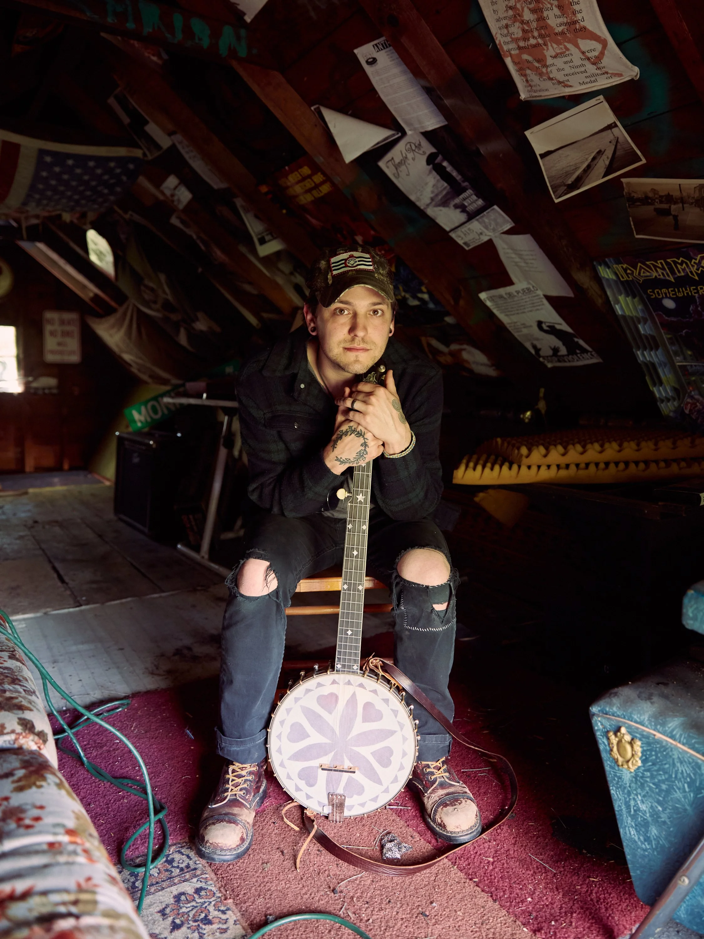A photograph of a person holding a banjo. The room has various posters, photographs, and musical equipment on the walls and floor.