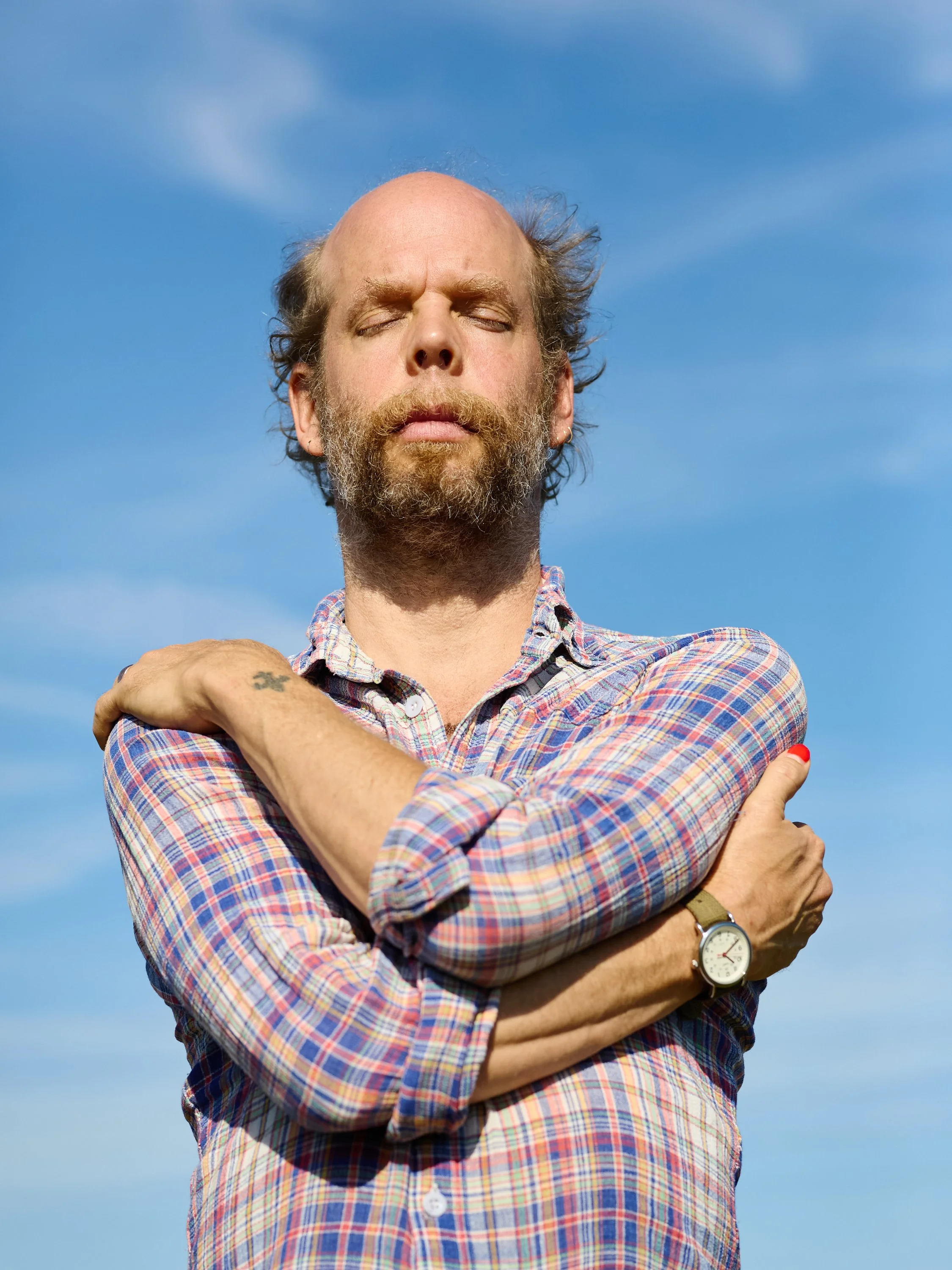 A person with closed eye, standing outdoors against a blue sky with some clouds, wearing a plaid shirt with rolled-up sleeves, a watch, and red nail polish.