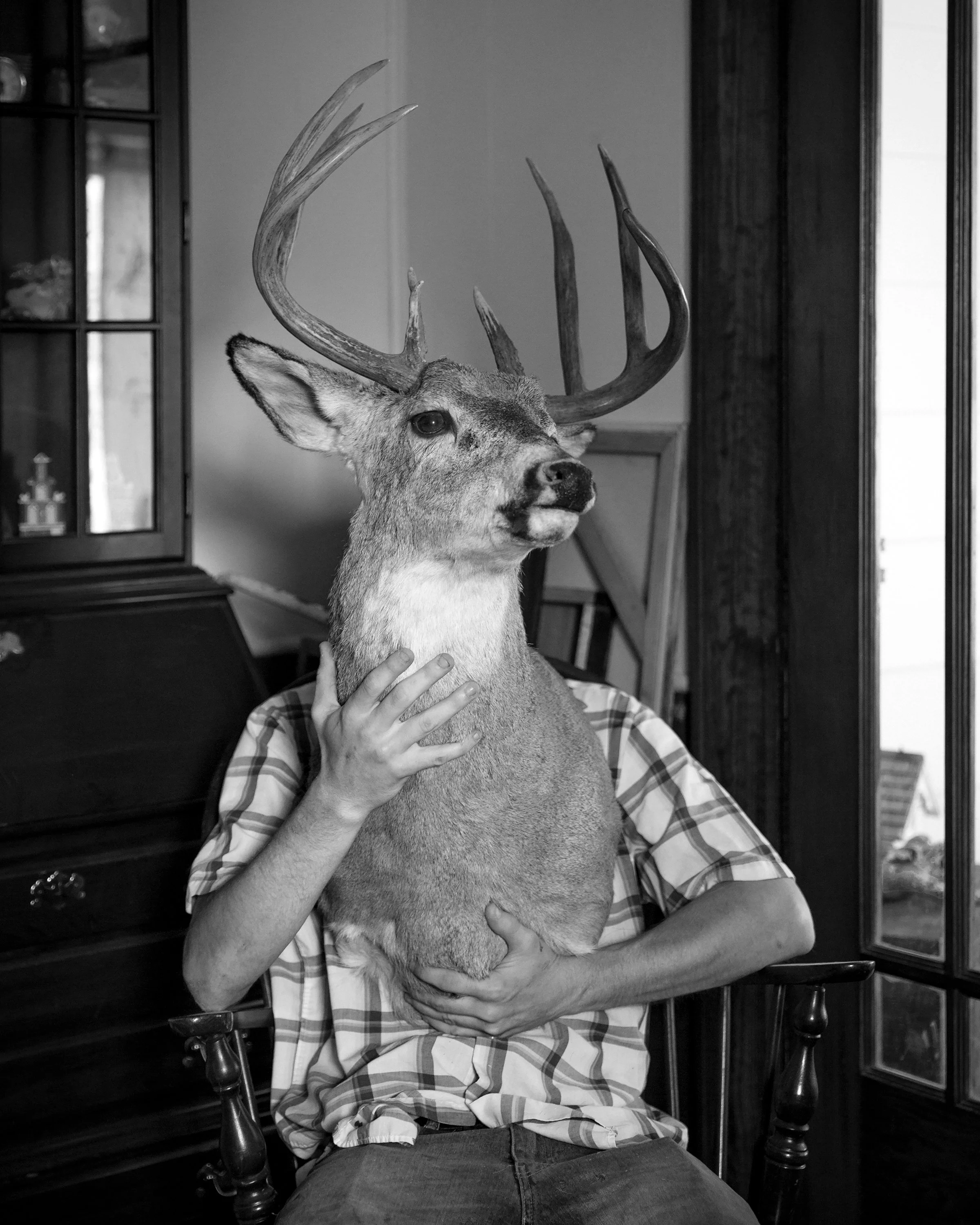 A black and white photo of a person sitting on a chair holding a deer's head with antlers, inside a room with a wooden cabinet and window.