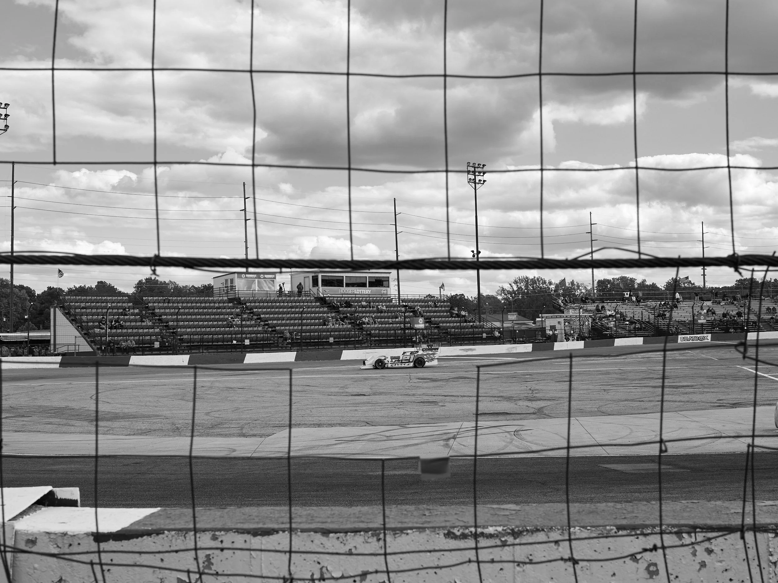 Black and white image of a race car on a racetrack, viewed through a fence with empty grandstands and cloudy sky in the background.
