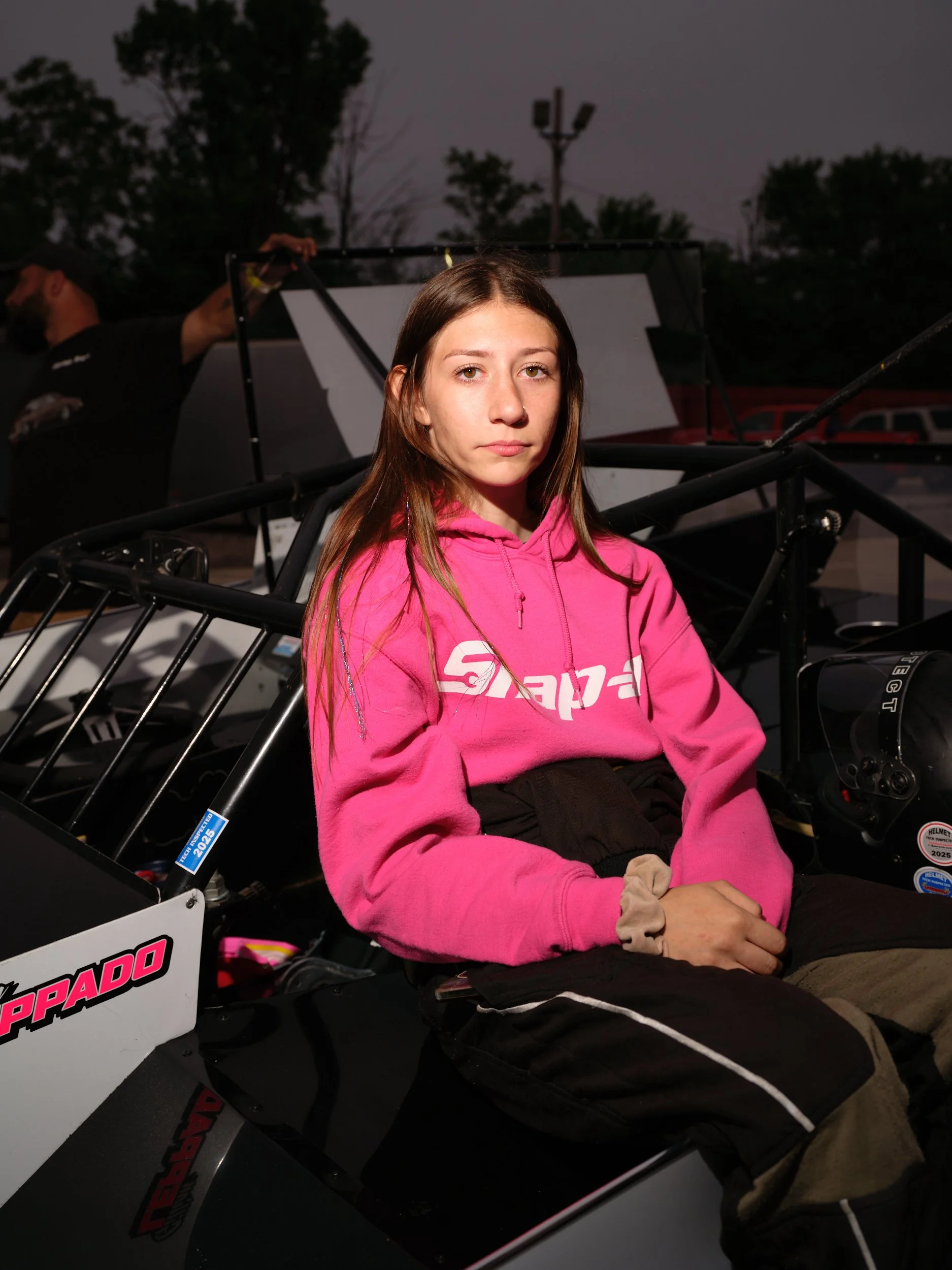 A young woman in a pink hoodie sitting in a race car, with a serious expression, outdoors during dusk.