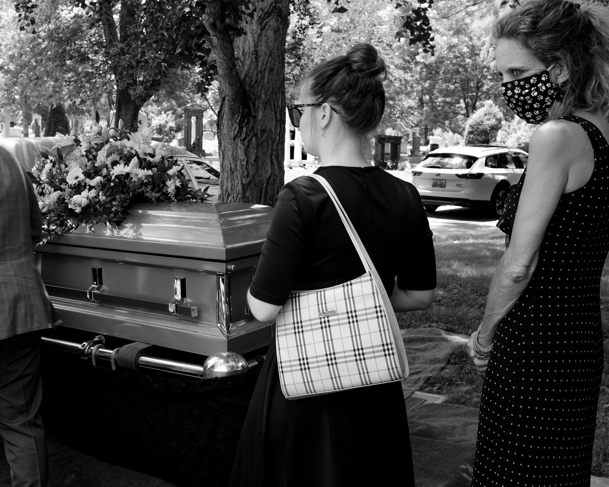 Two women wearing masks stand near a funeral casket with a floral arrangement outdoors, with cars and trees in the background.