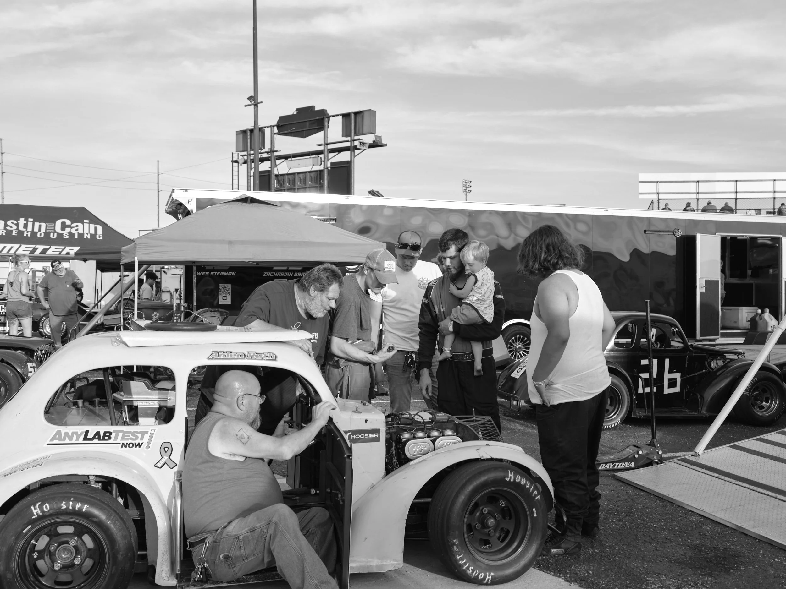 People gather around a race car in a paddock area at a motorsport event, with team members working on the car and others observing.