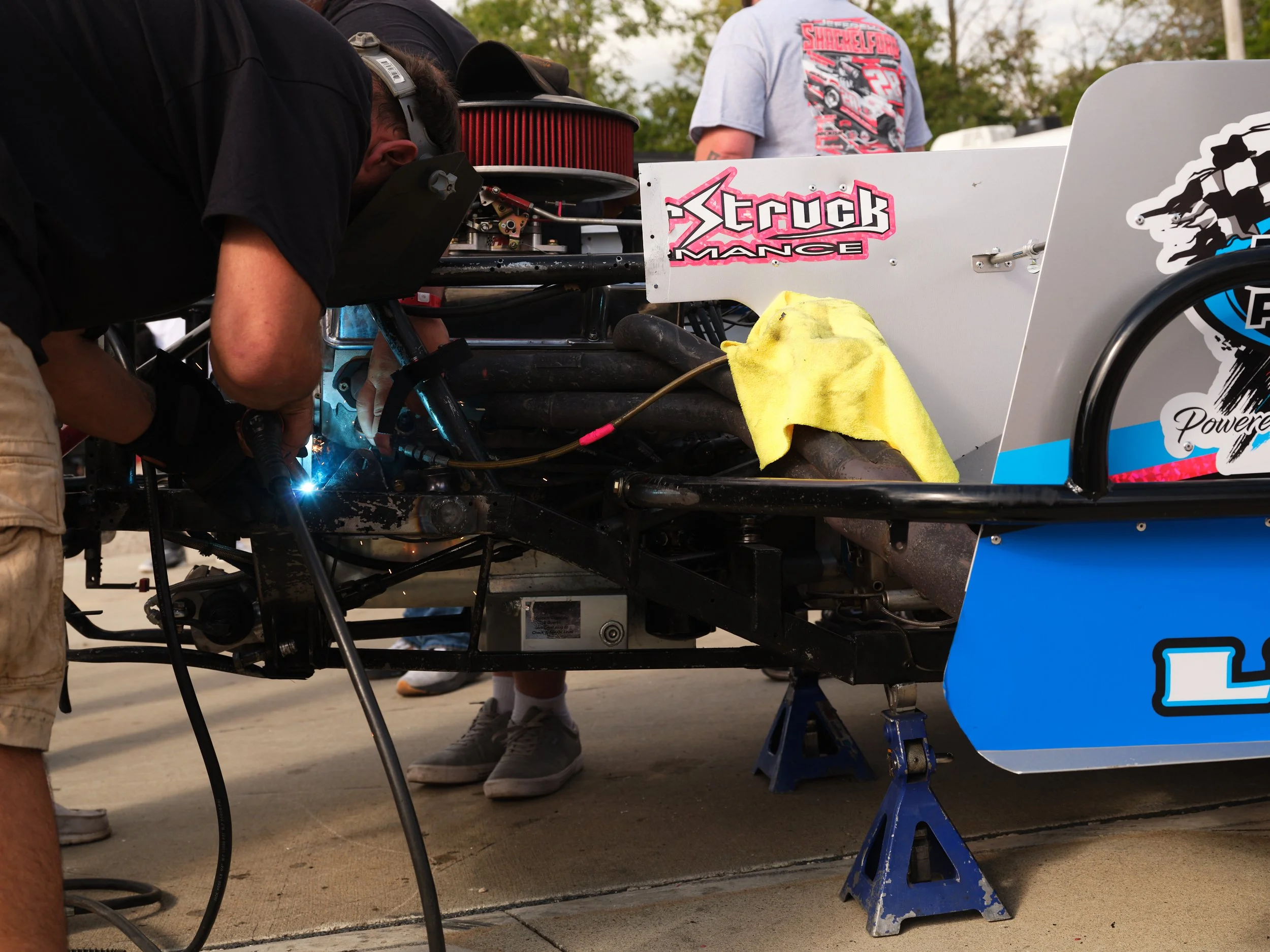 A mechanic welding or repairing a race car, with two people visible nearby, one in a gray t-shirt and the other partially visible. The race car has sponsor logos and is elevated on jack stands.