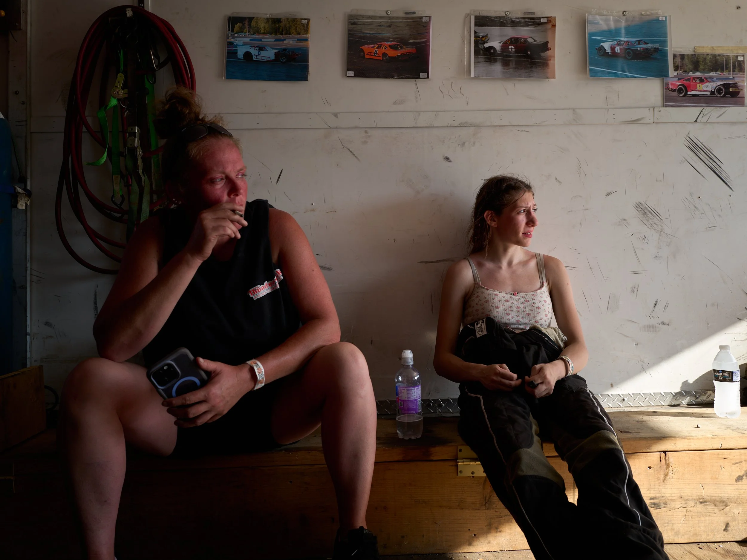 Two women sitting on a wooden bench in a garage, with race car photos on the wall above them, one holding a mobile phone, the other holding some clothing, both looking away from the camera.